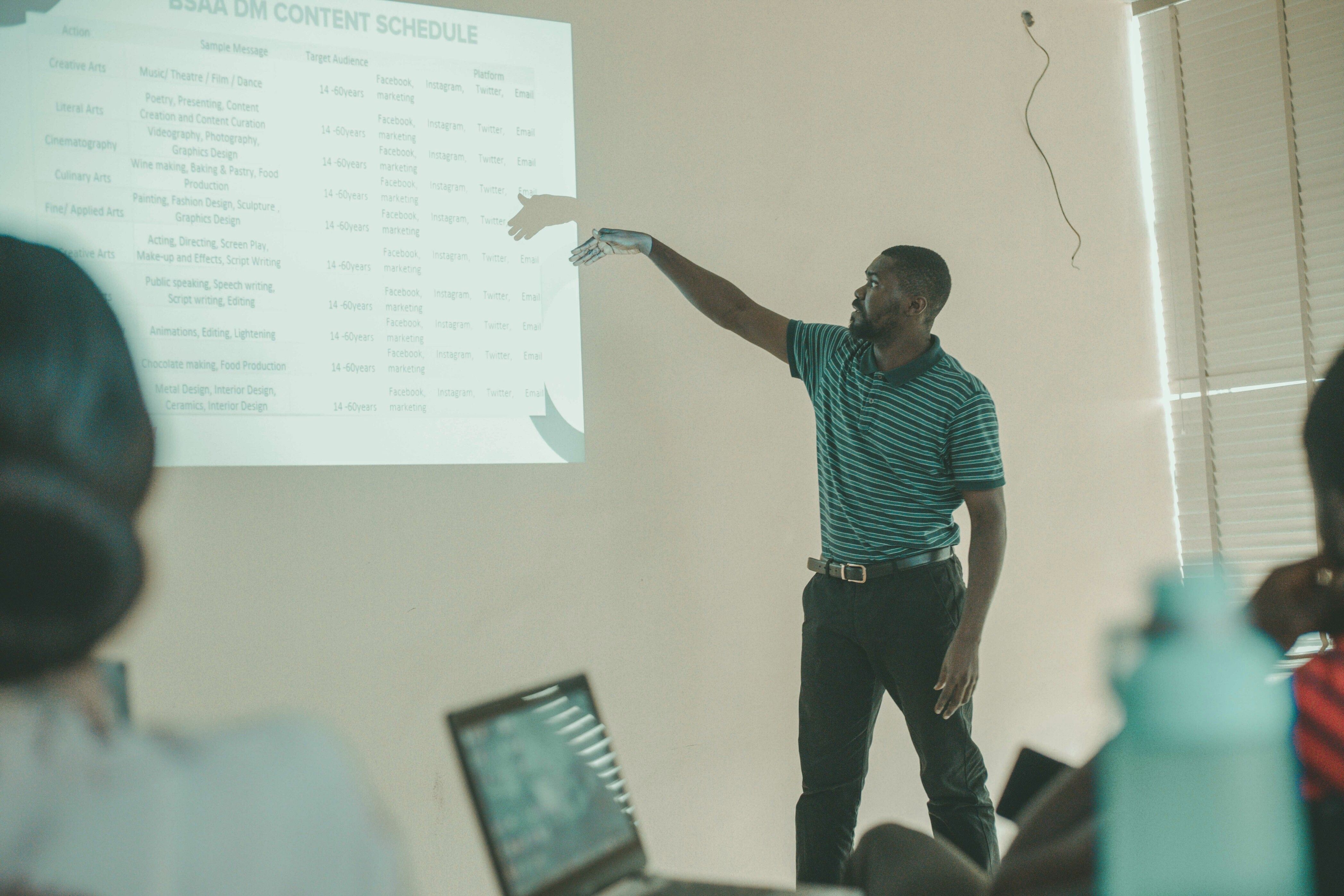 man standing in front of a classroom and pointing toward a projected presentation slide showing a content schedule.
