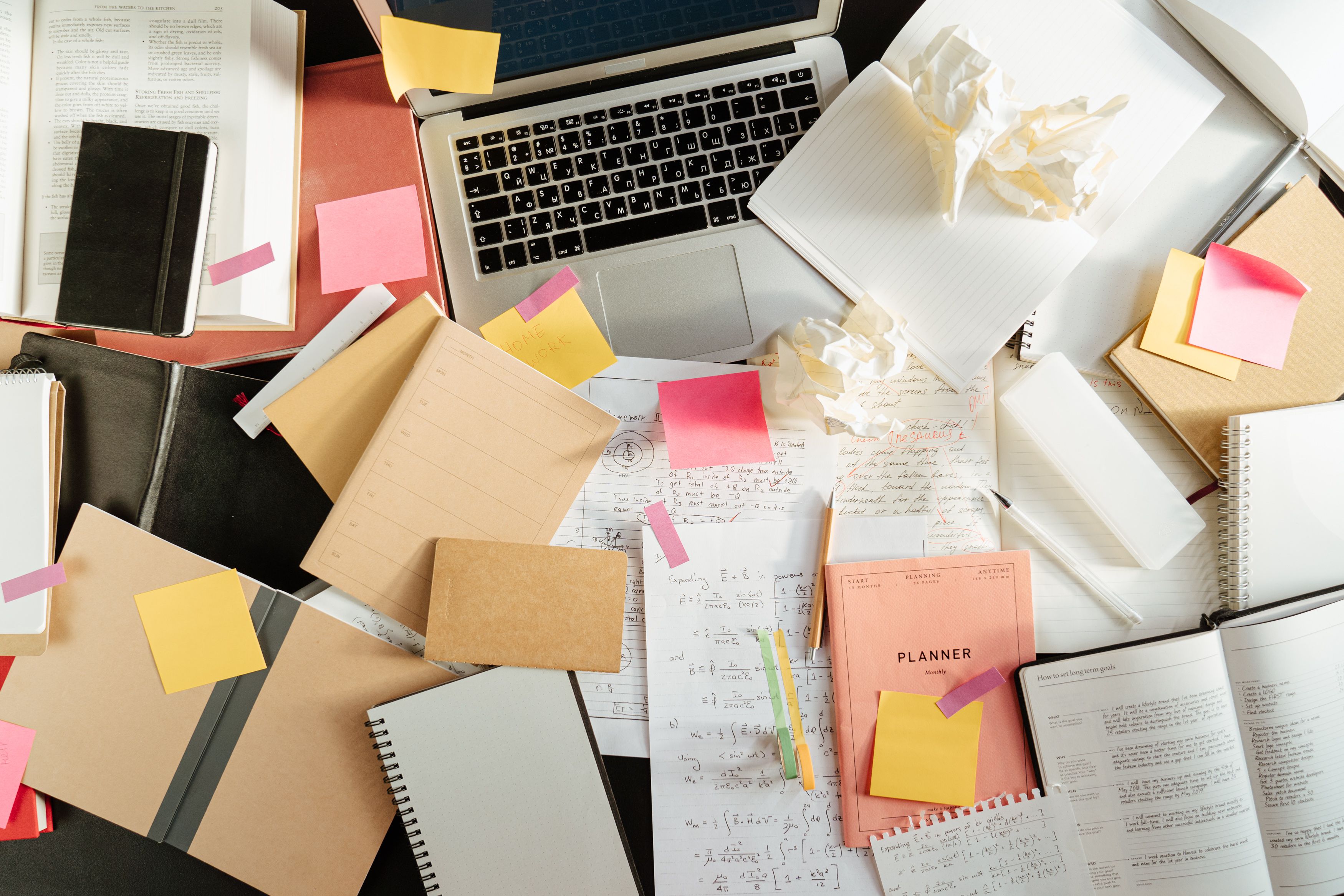 A cluttered office desk covered with open notebooks, sticky notes, handwritten math equations, and an open laptop.