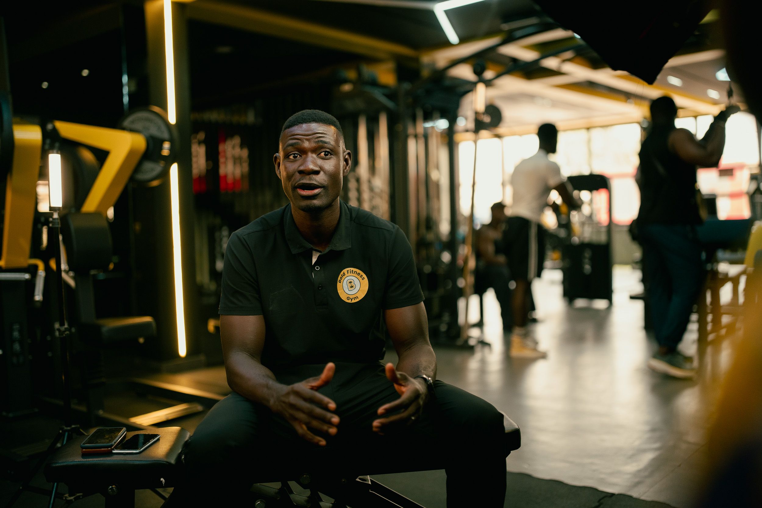 A fitness trainer sitting on an exercise bench inside a gym and gesturing while speaking in an interview setup.