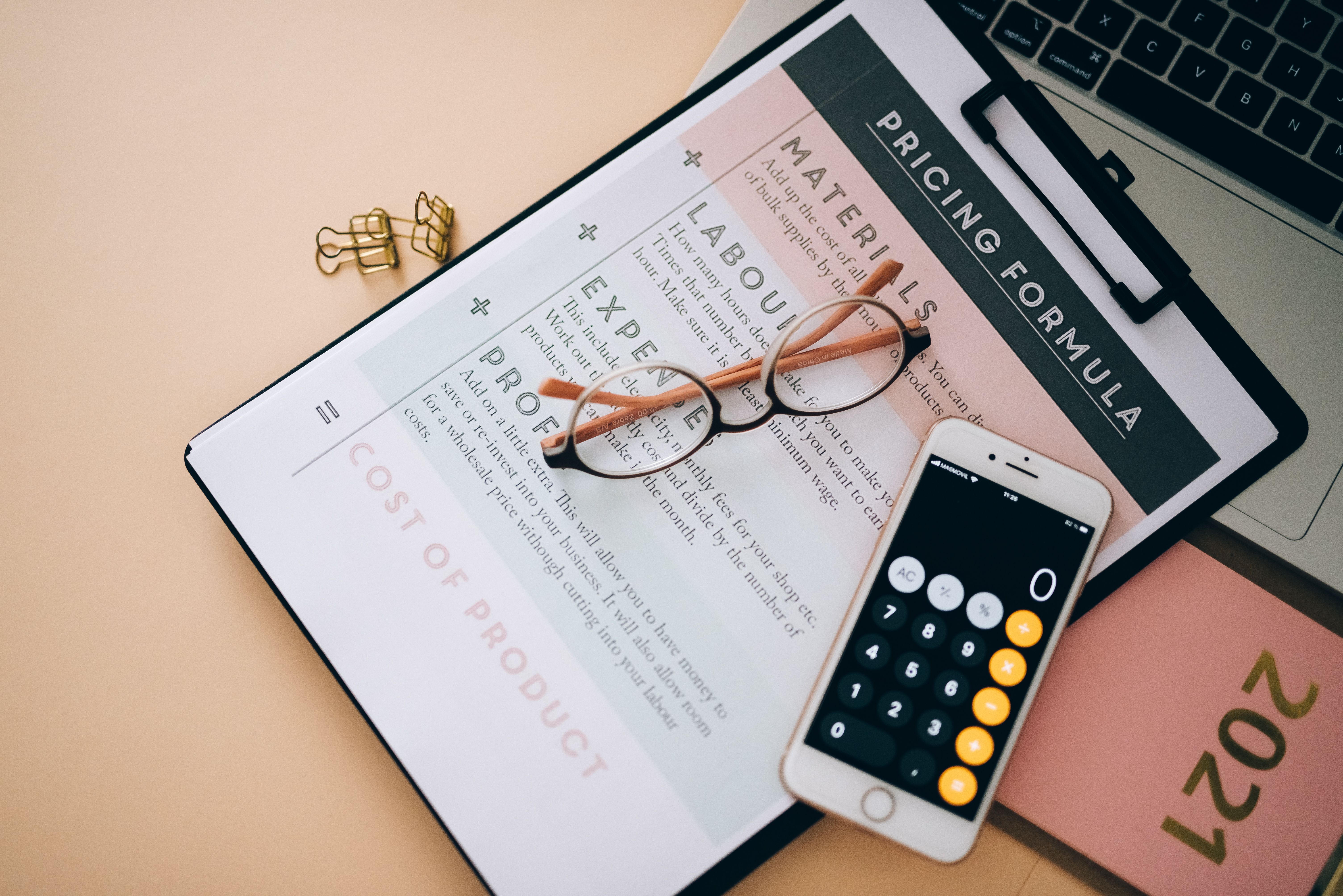 A clipboard displaying a pricing formula document next to a smartphone showing a calculator screen and eyeglasses.
