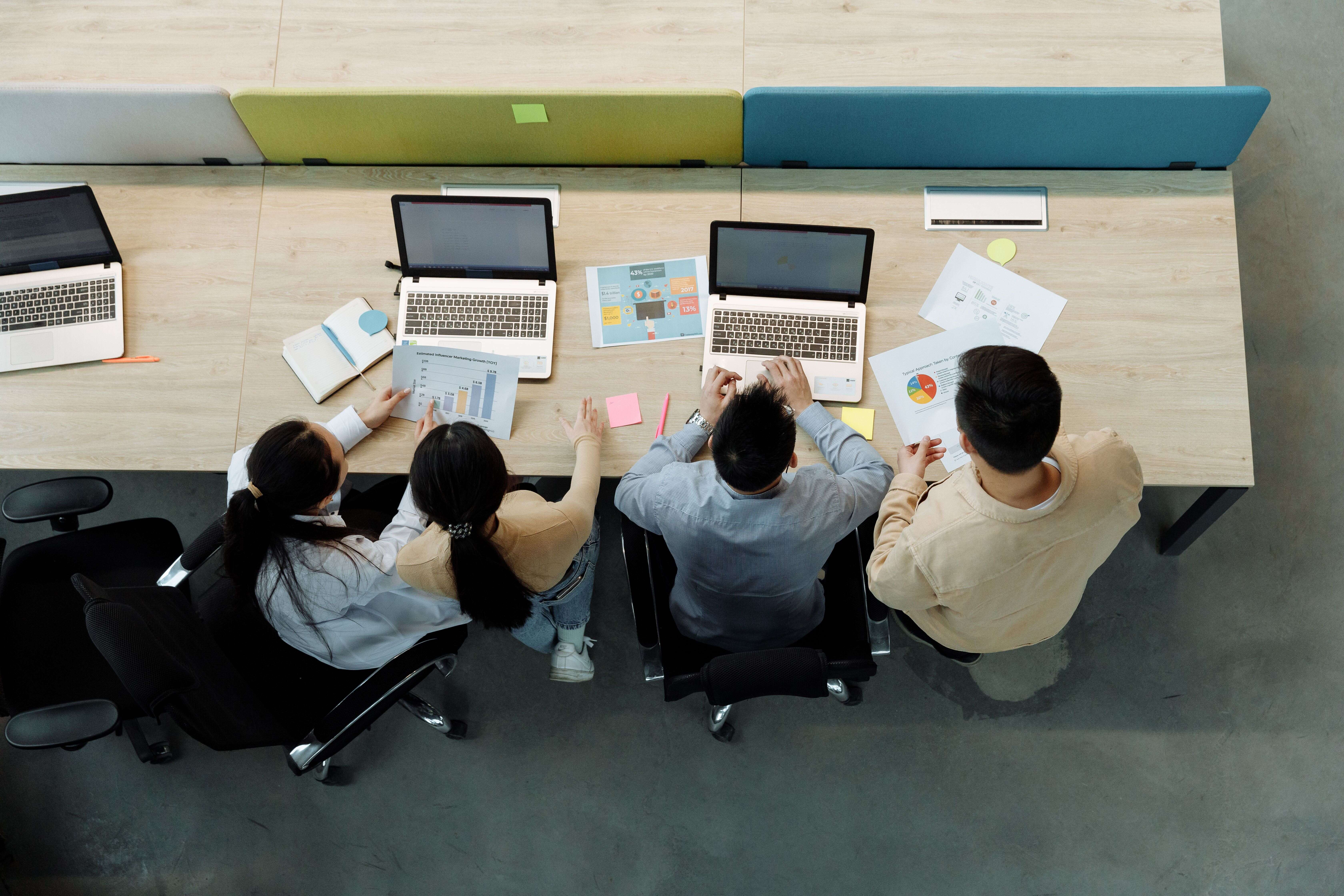 Top-down view of four colleagues sitting together at a shared wooden office table working on laptops and analyzing chart reports.