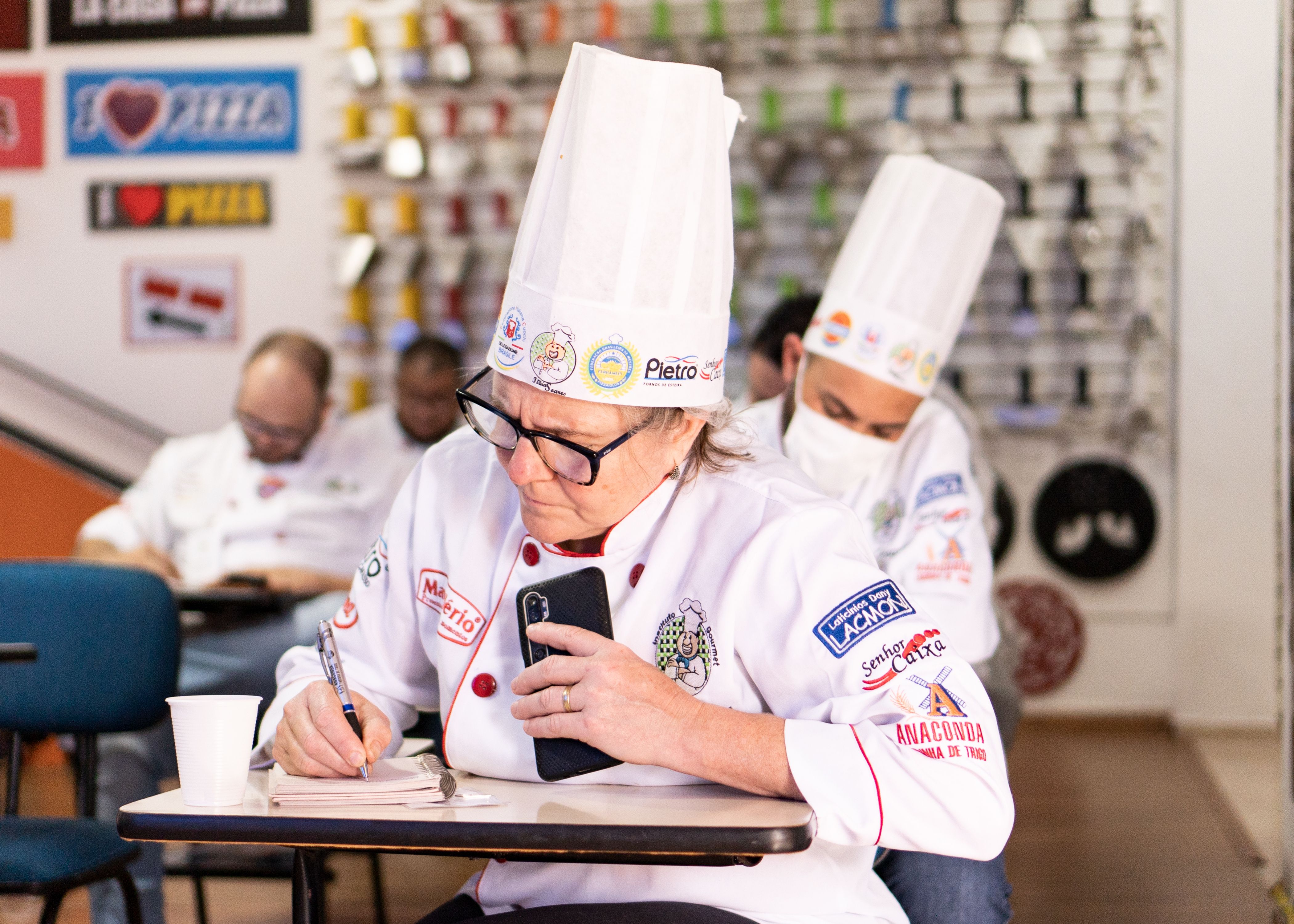 A chef in a white toque uniform taking notes in a notepad with a pen during a training session.