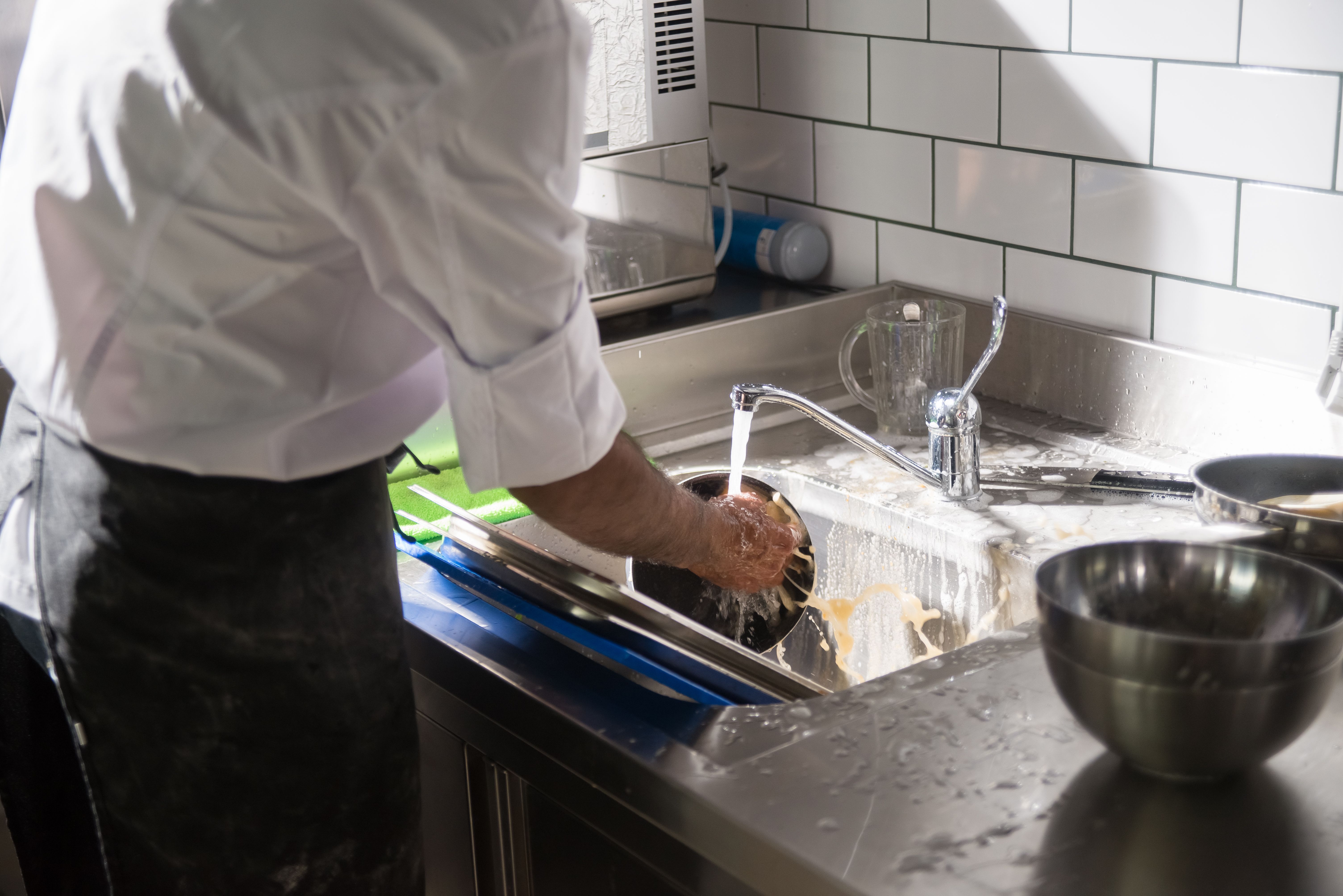 A kitchen staff member washing stainless steel cooking pots in a deep commercial sink filled with soapy water.