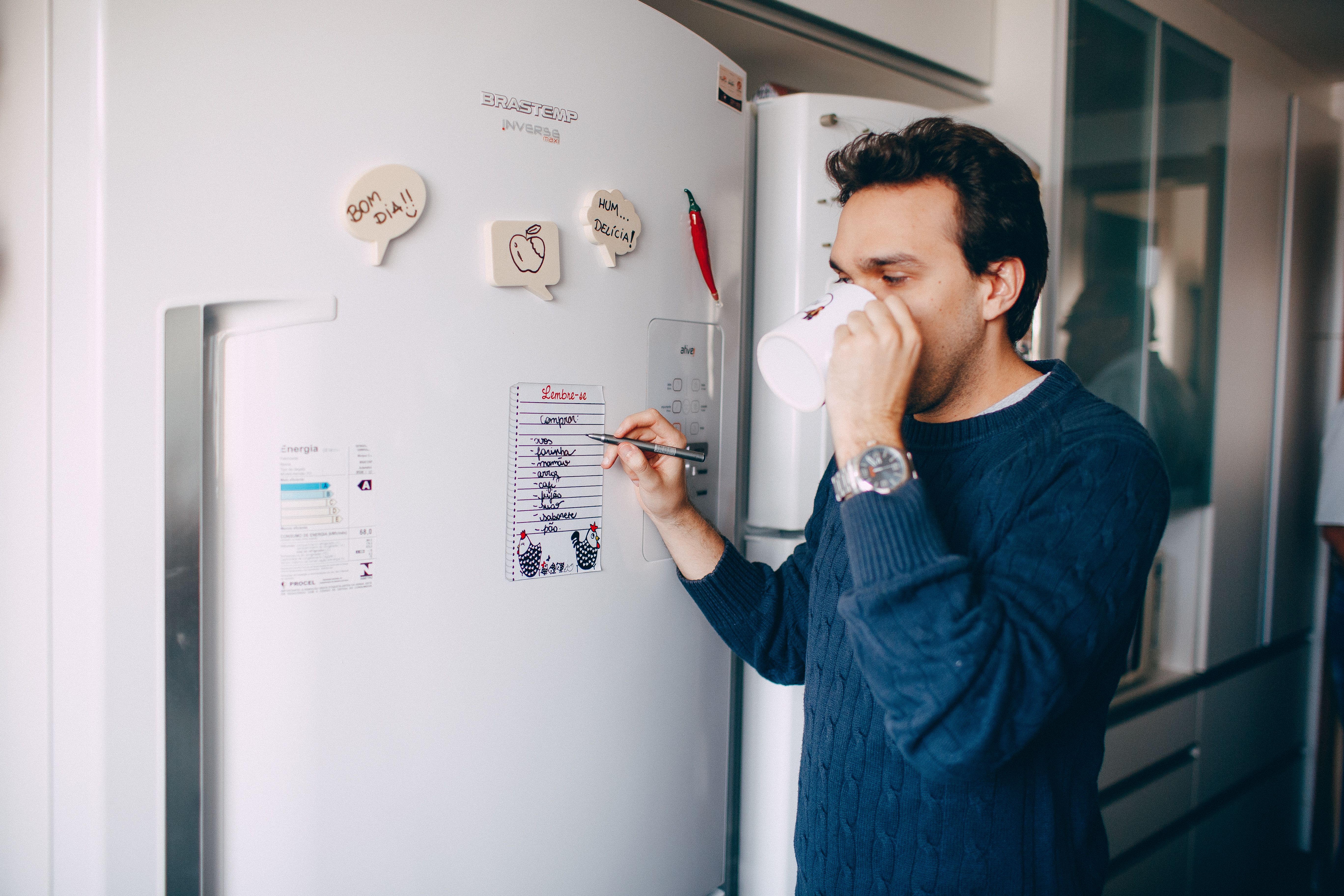A man drinking from a mug in a kitchen while writing a handwritten list on a note attached to a refrigerator.