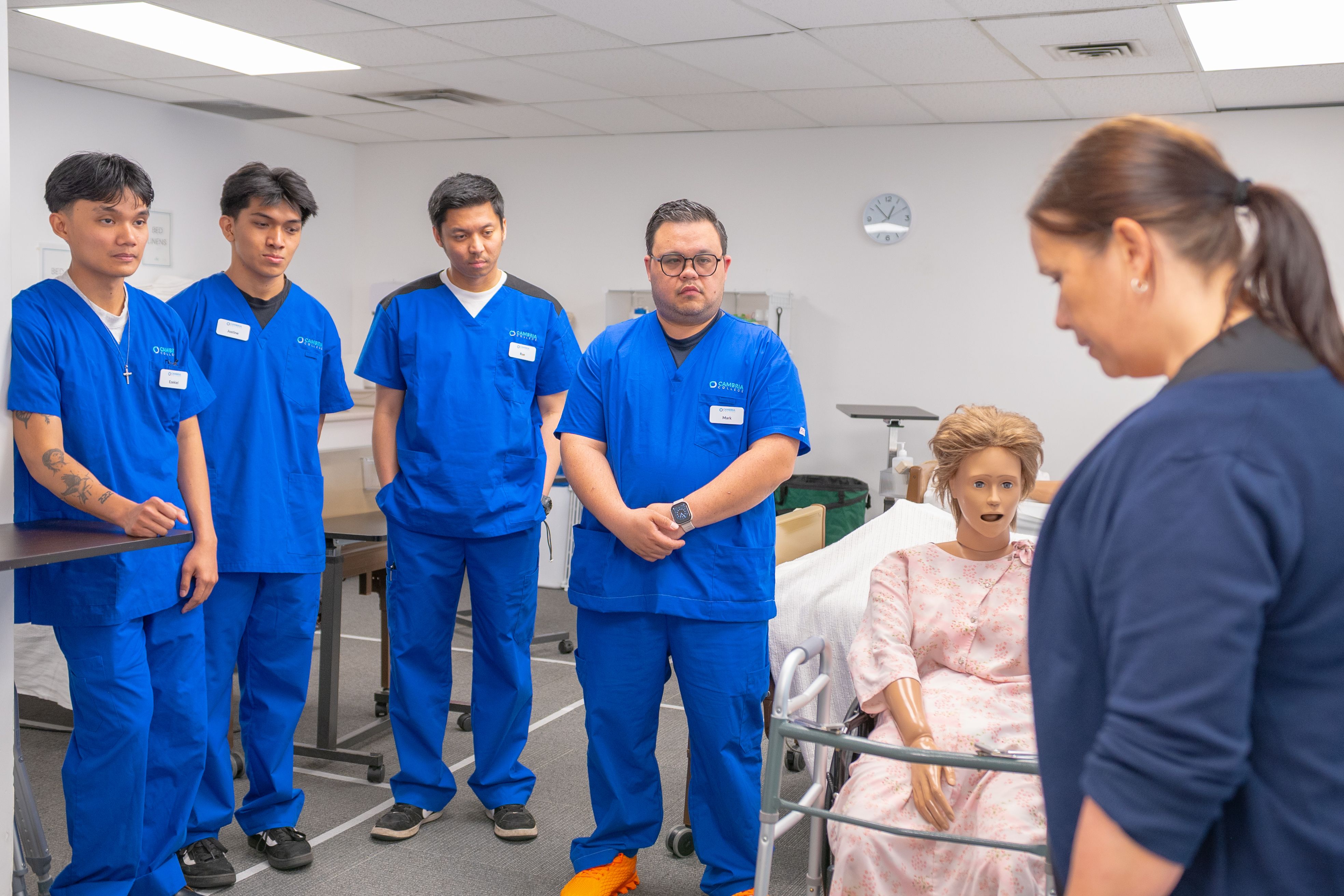 A group of nursing students in blue scrubs listening attentively during a medical training session with a mannequin.