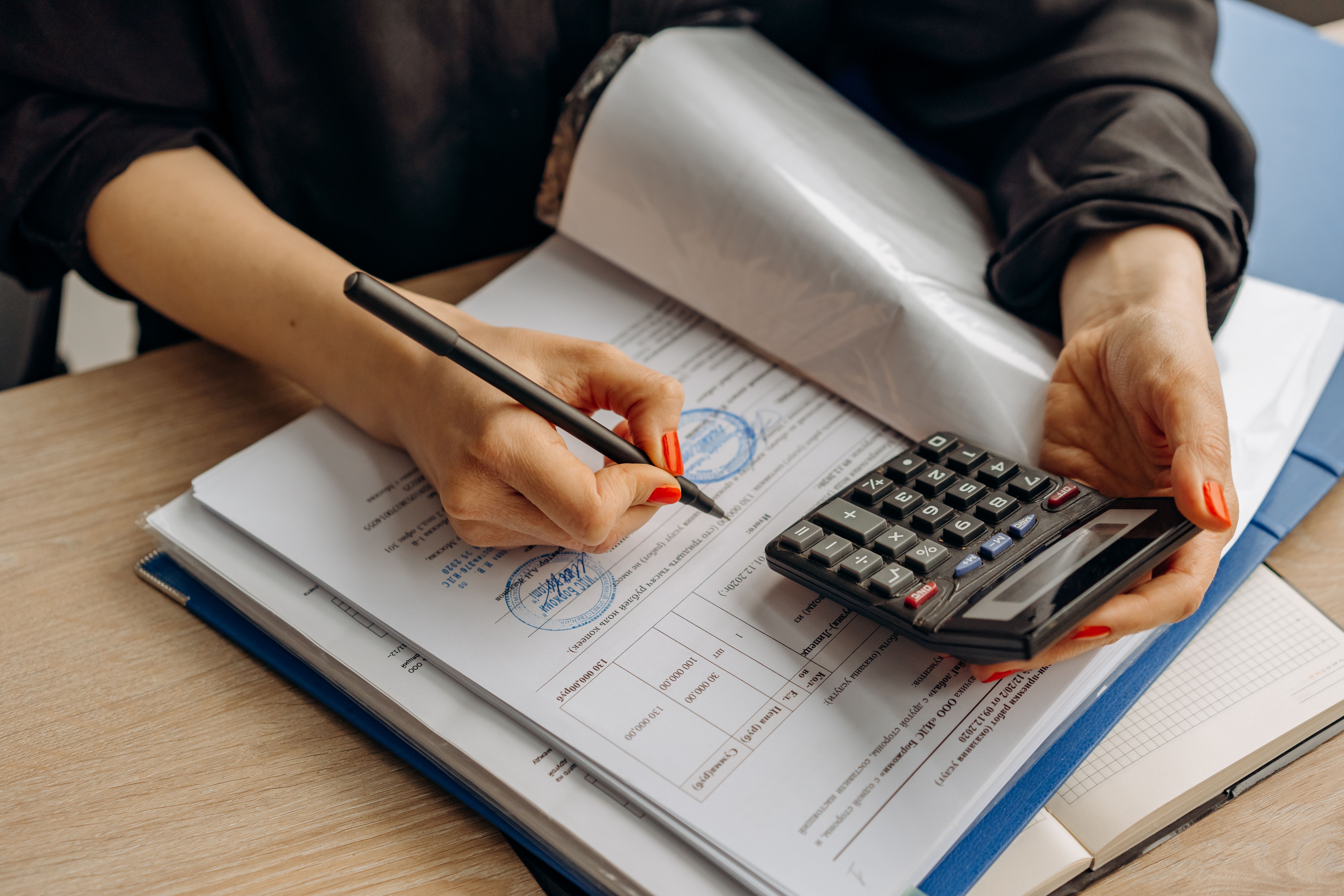 Close-up of a person using a calculator with one hand while holding a pen to fill out official paperwork.
