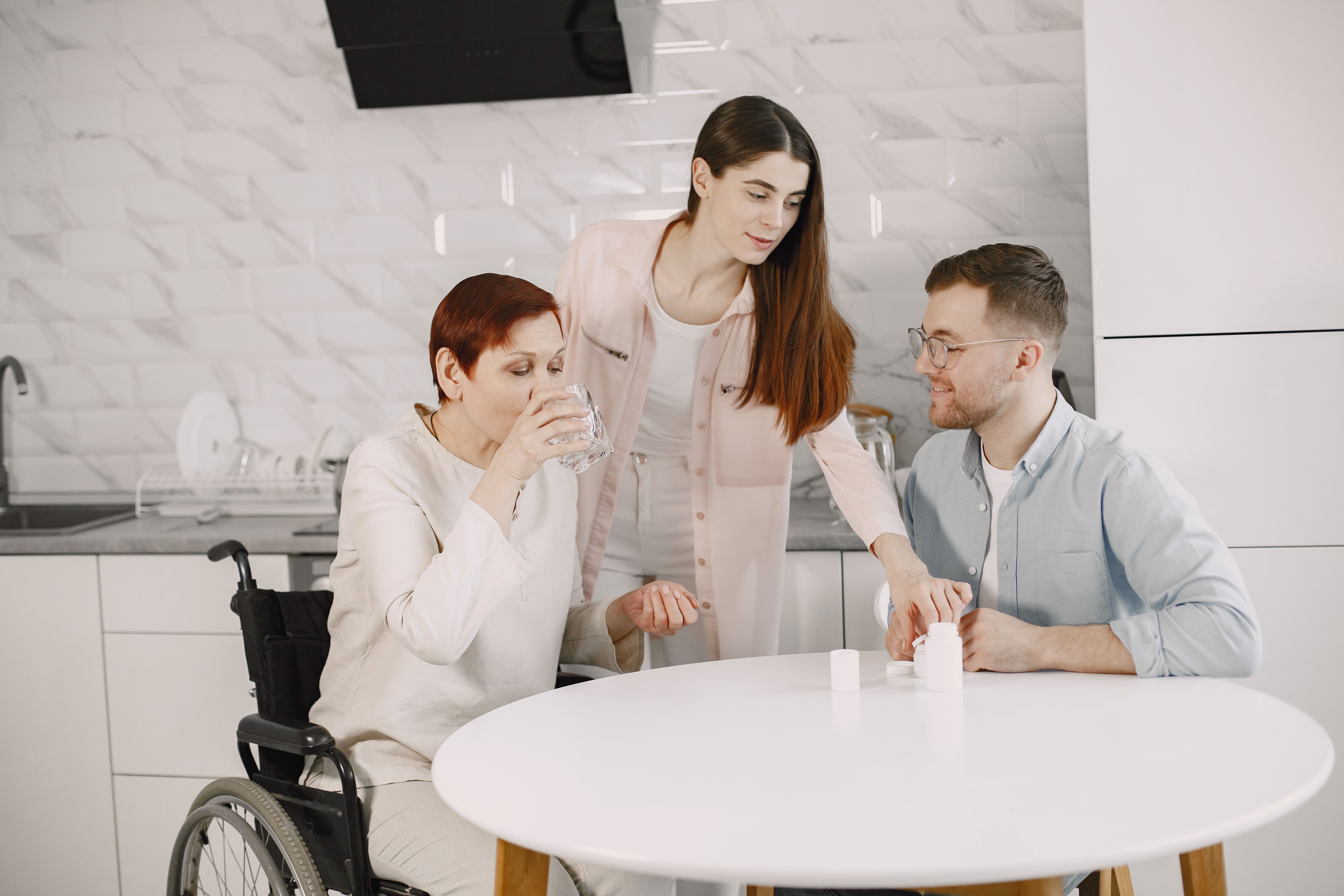 A woman in a wheelchair drinking water at a kitchen table while caregivers assist her with medication bottles.