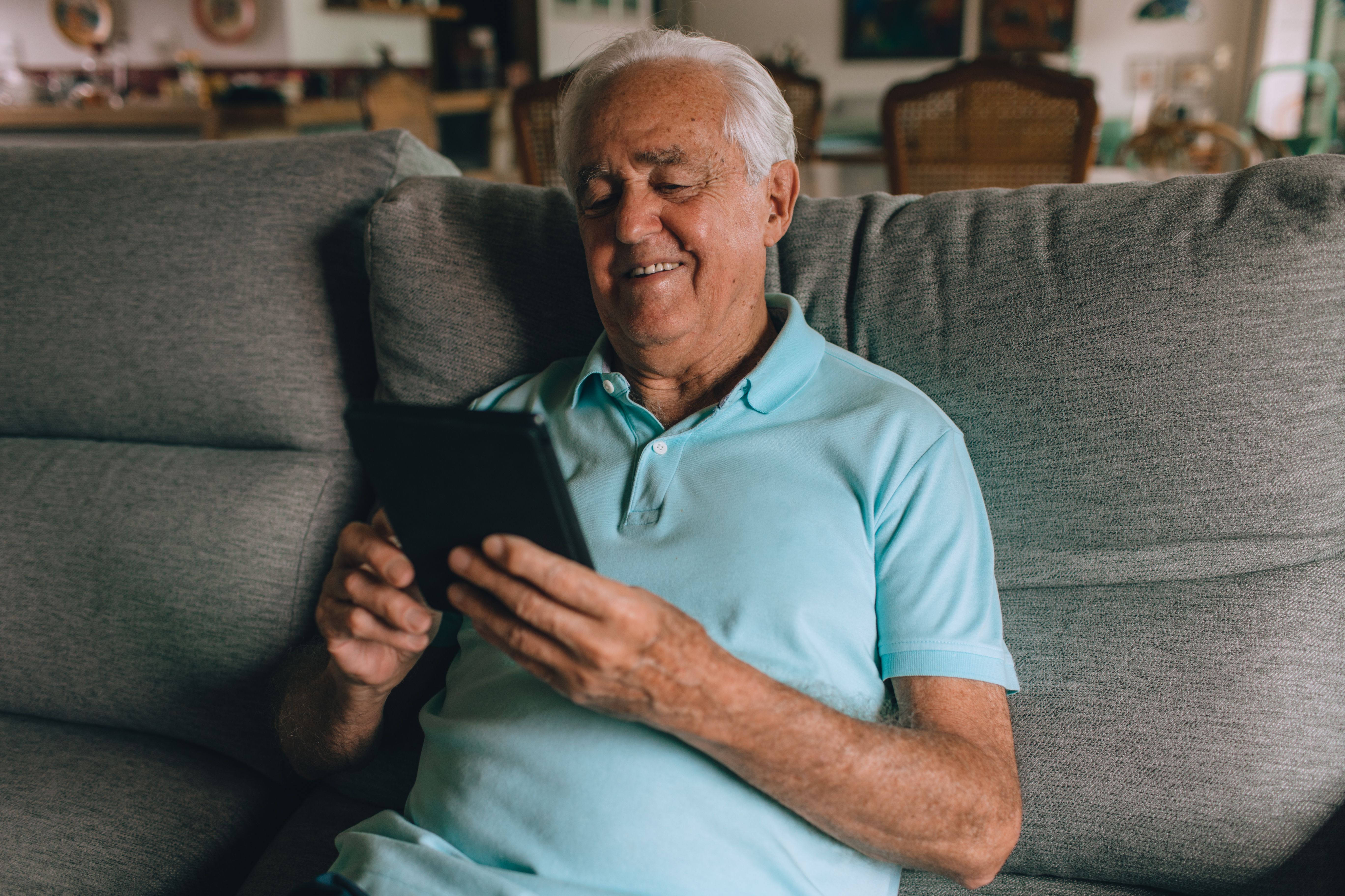 An elderly man smiling while sitting on a grey sofa and holding a digital tablet device.
