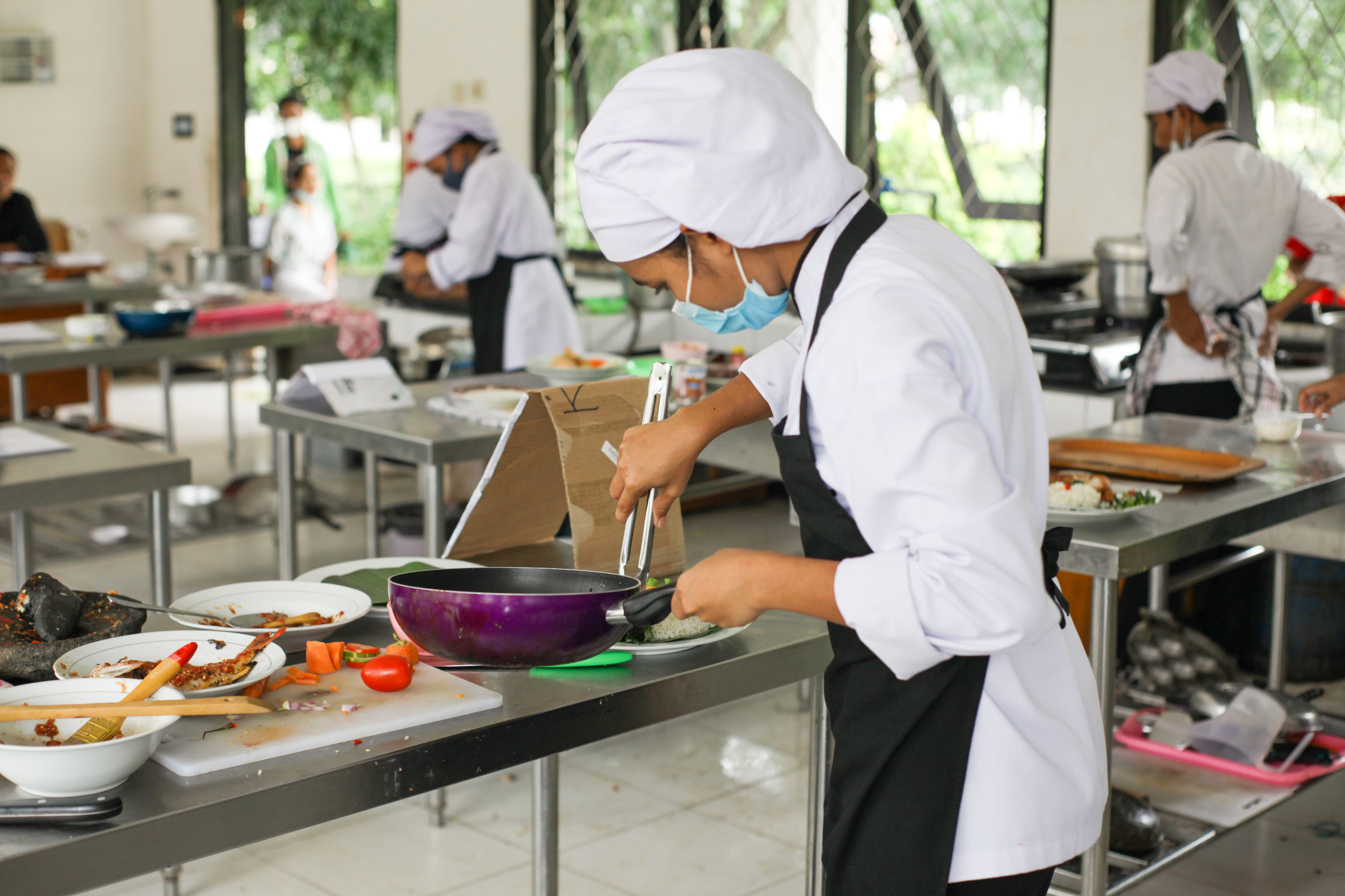 A young culinary student wearing a chef toque and face mask using metal tongs to cook food in a frying pan during a training class.