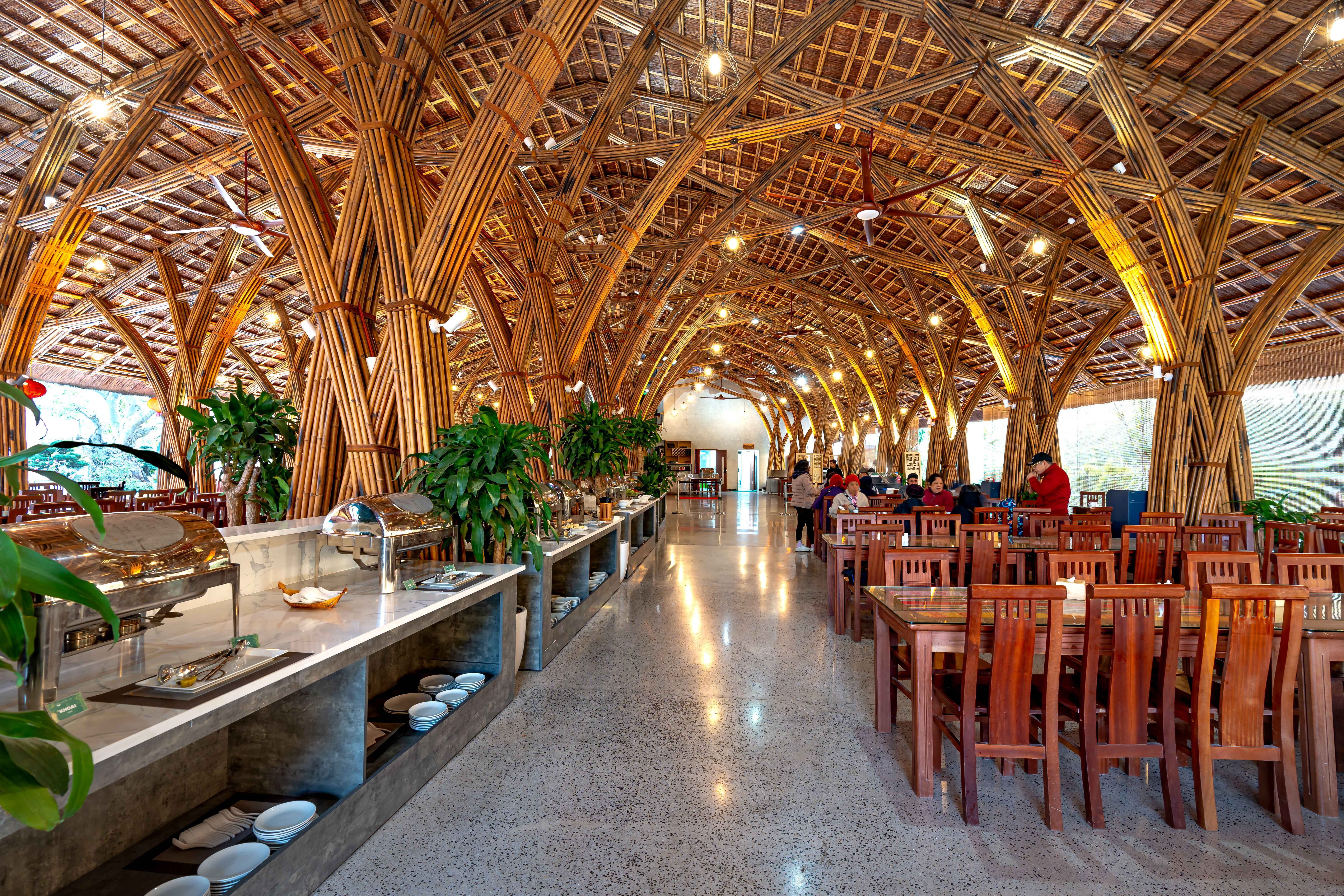 A spacious resort dining hall featuring an arched bamboo ceiling structure, wooden tables, and a breakfast buffet setup.