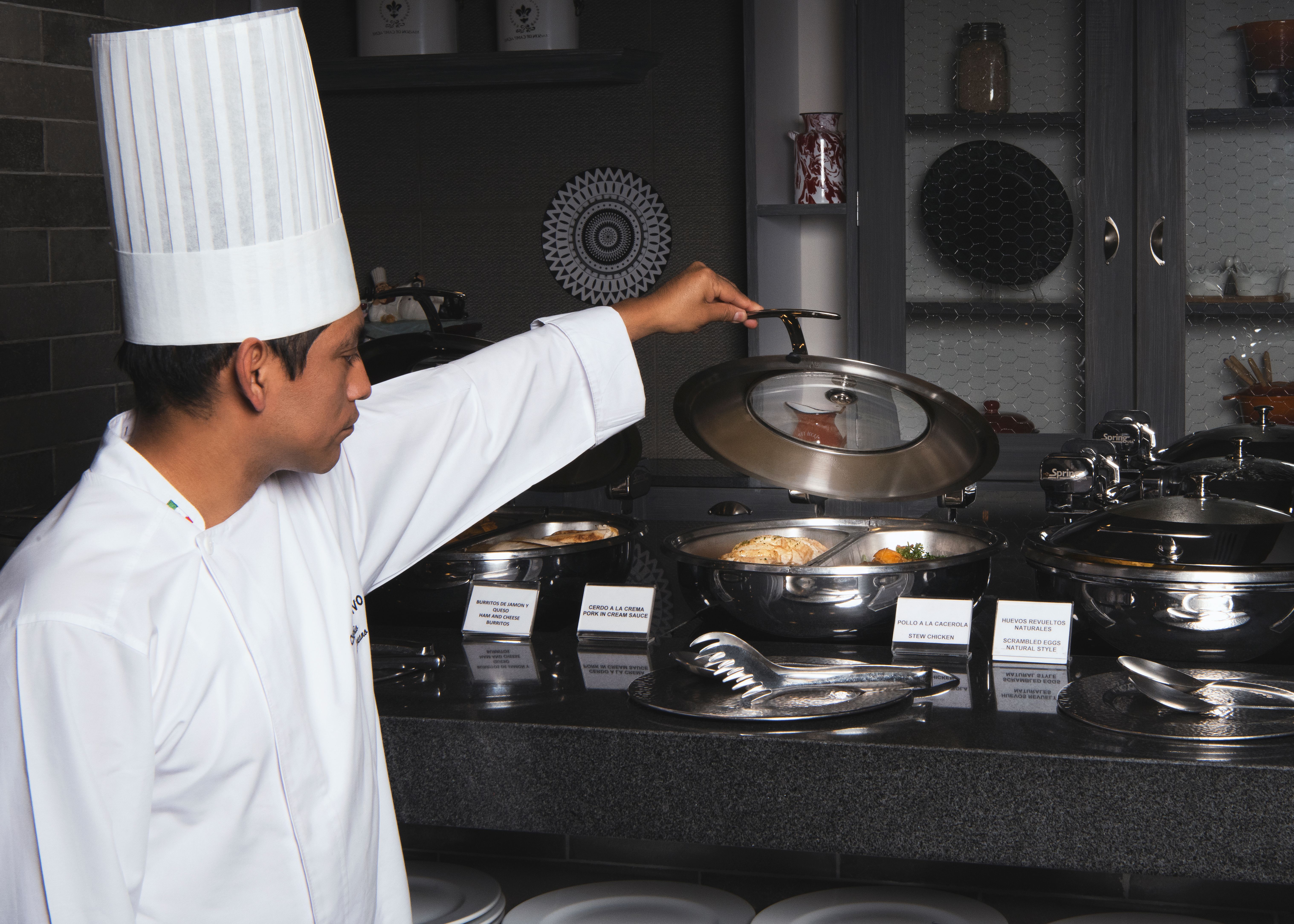 A chef lifting a metal lid off a chafing dish on a buffet counter to inspect hot food.