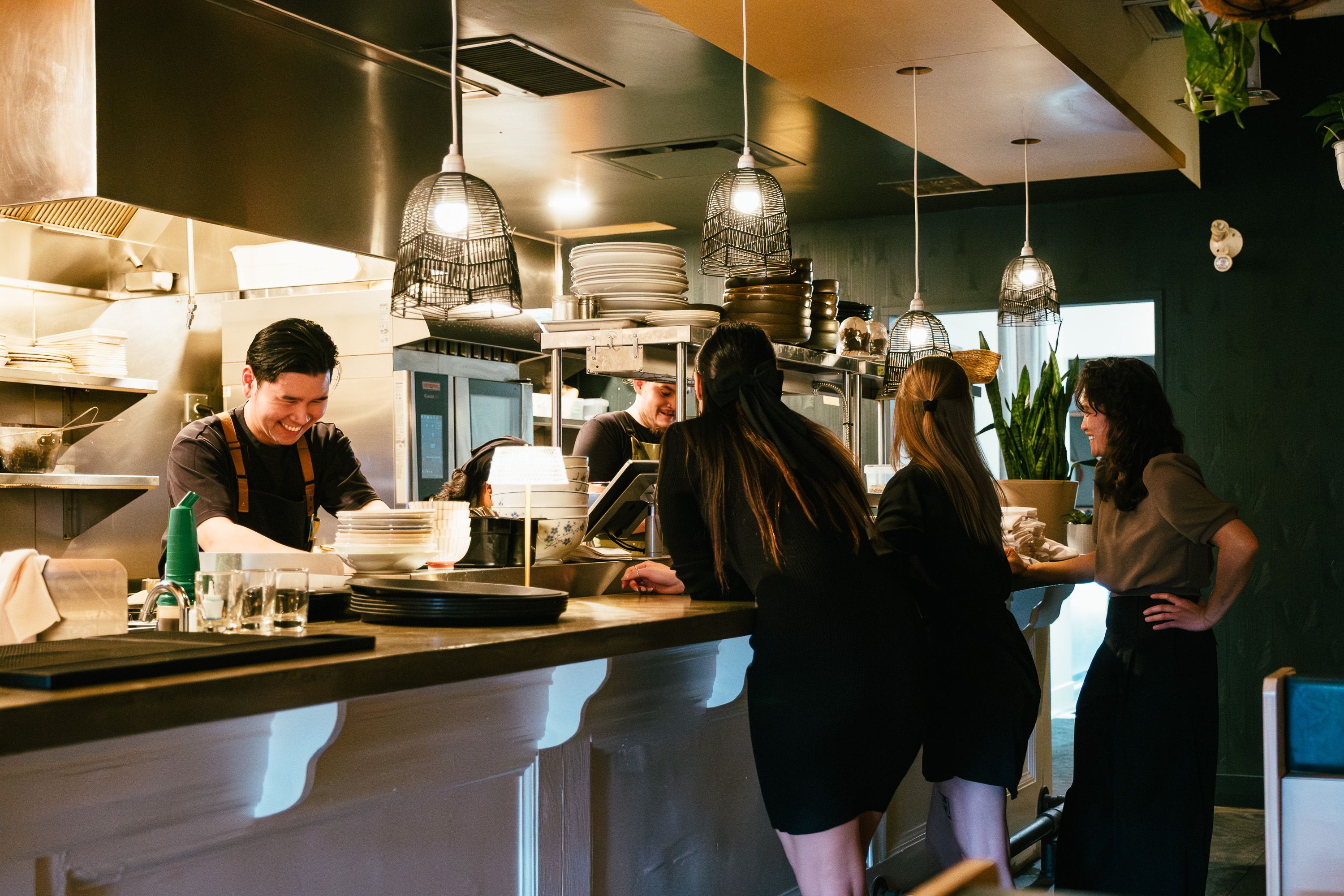 A smiling chef interacting with staff over a kitchen counter in a busy restaurant environment.