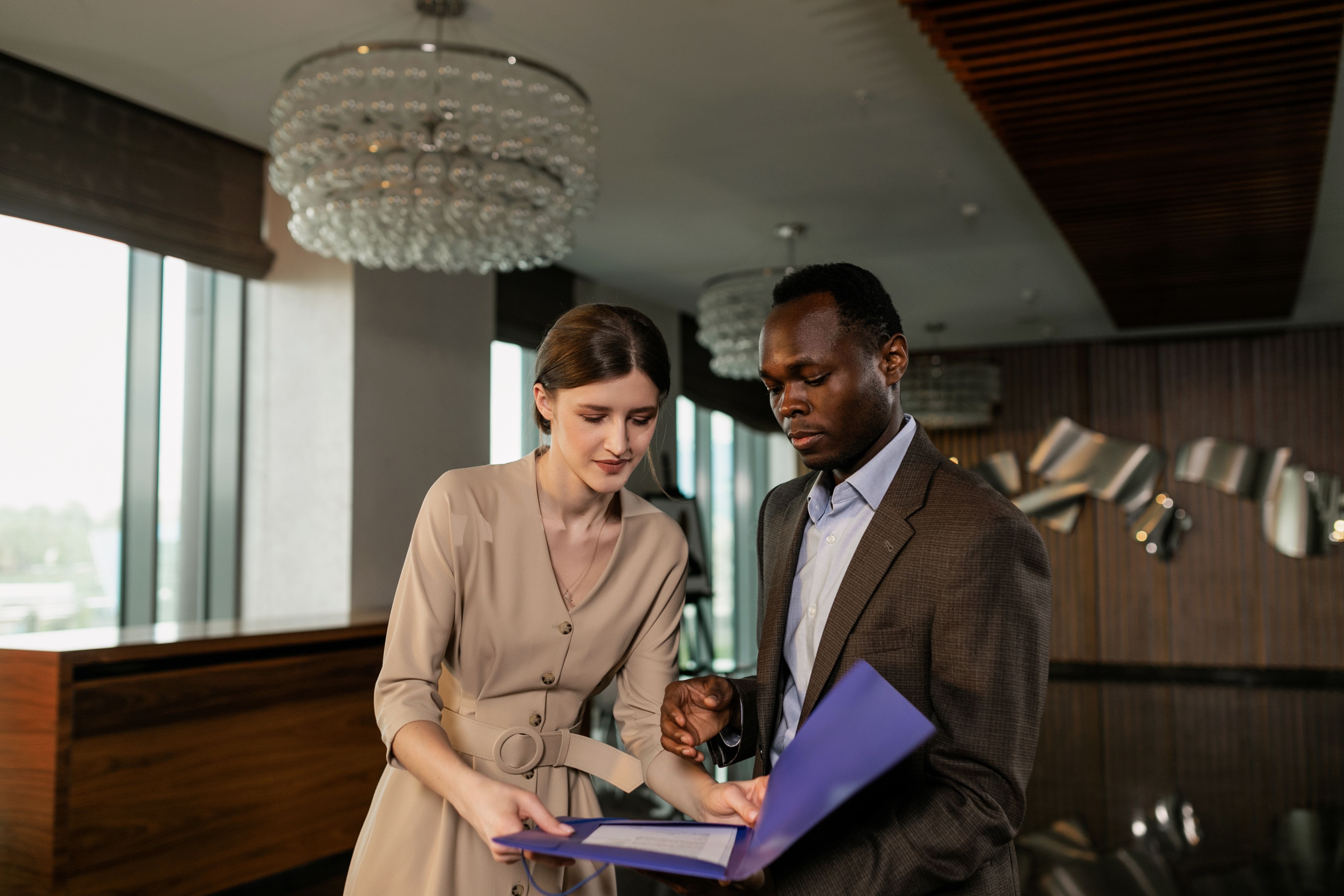 Two business professionals in a hotel lobby reviewing documents held inside a open blue folder.