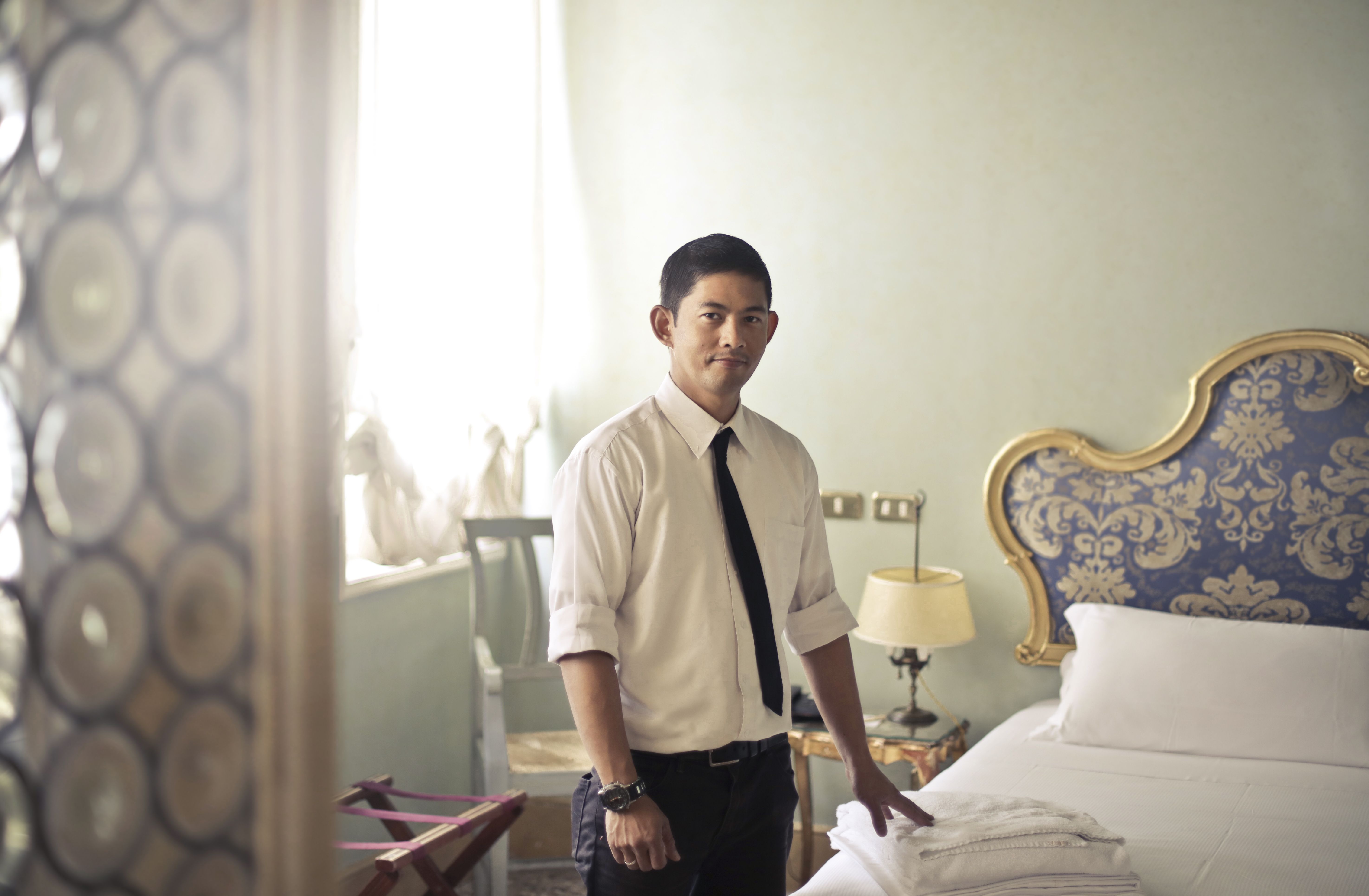 A hotel staff member in a white shirt and black tie placing fresh white towels on a bed in a guest room