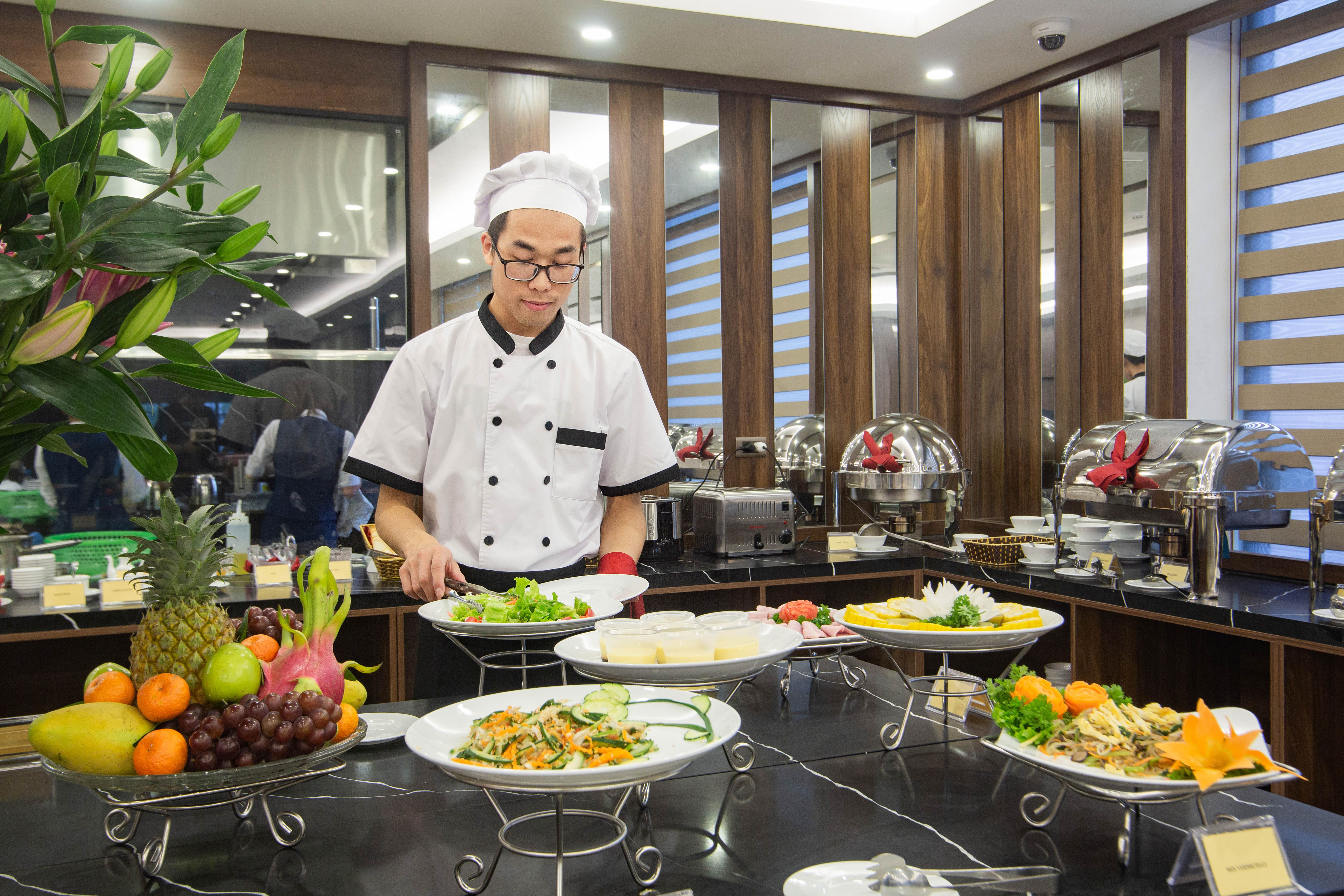 A chef in a white uniform arranging fresh salads and dishes on a hotel breakfast buffet line.