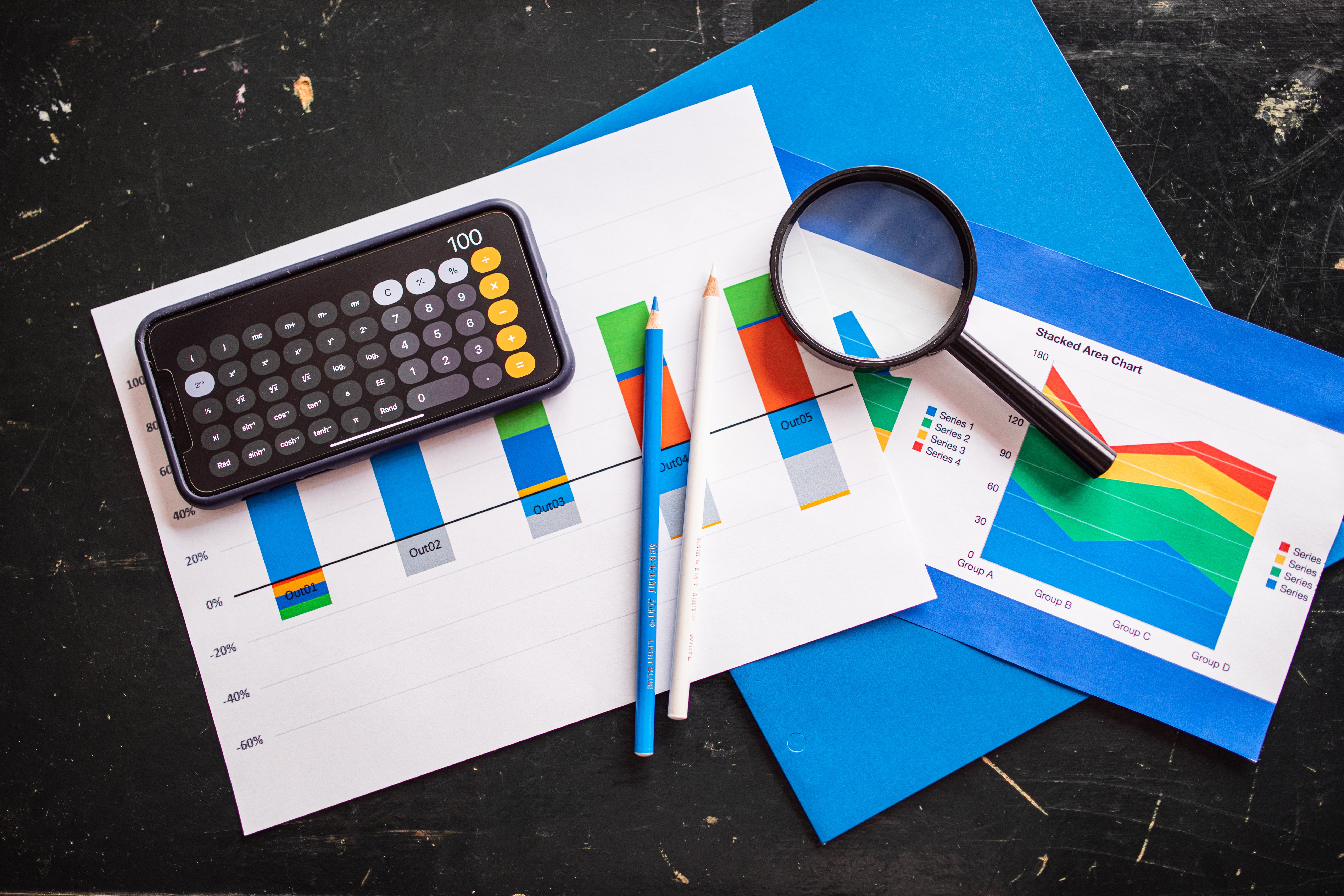 A magnifying glass, smartphone with a calculator app open, colored pencils, and printed data charts lying on a desk.