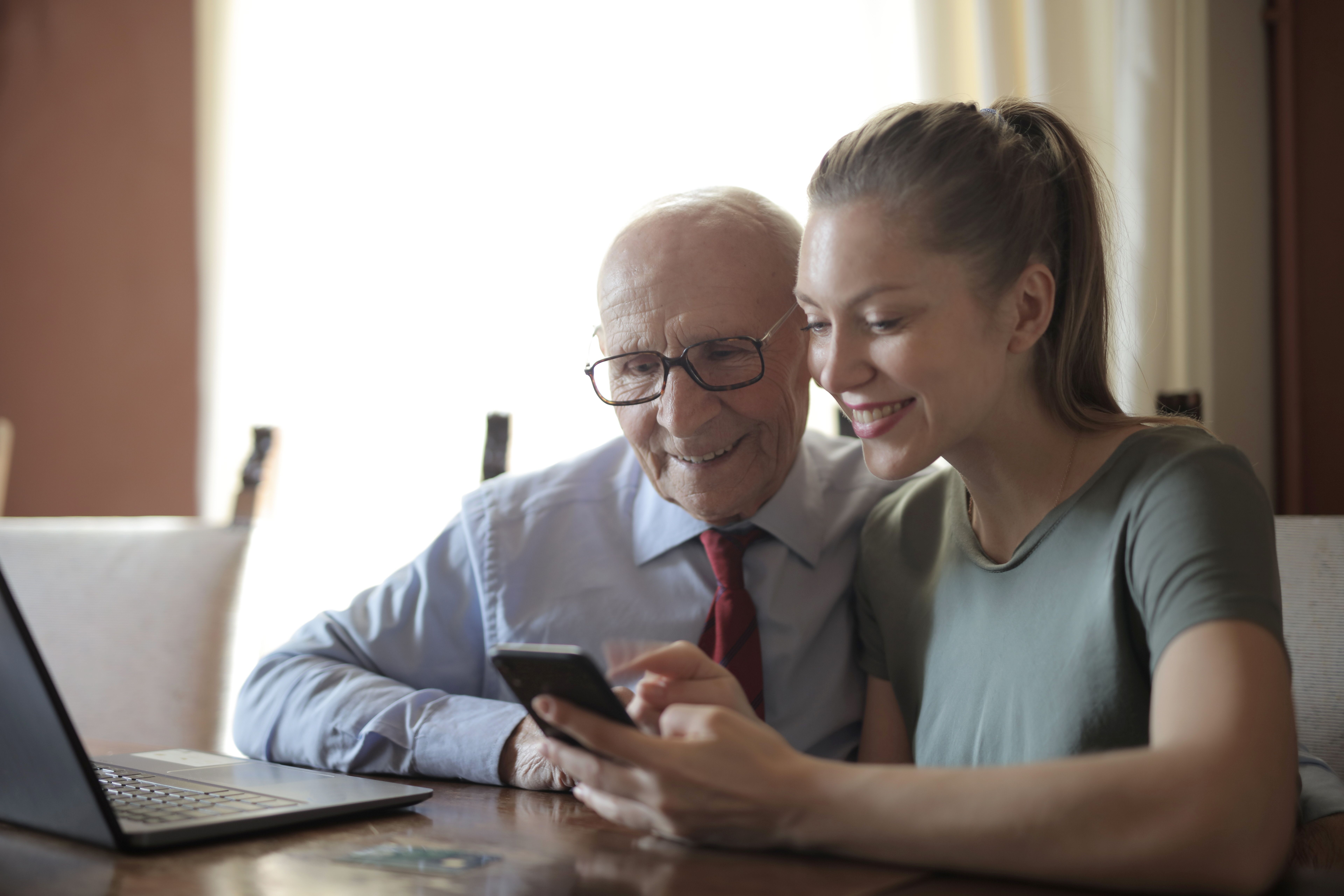 A young woman smiling while showing a smartphone screen to an elderly man wearing glasses and a tie at a table.
