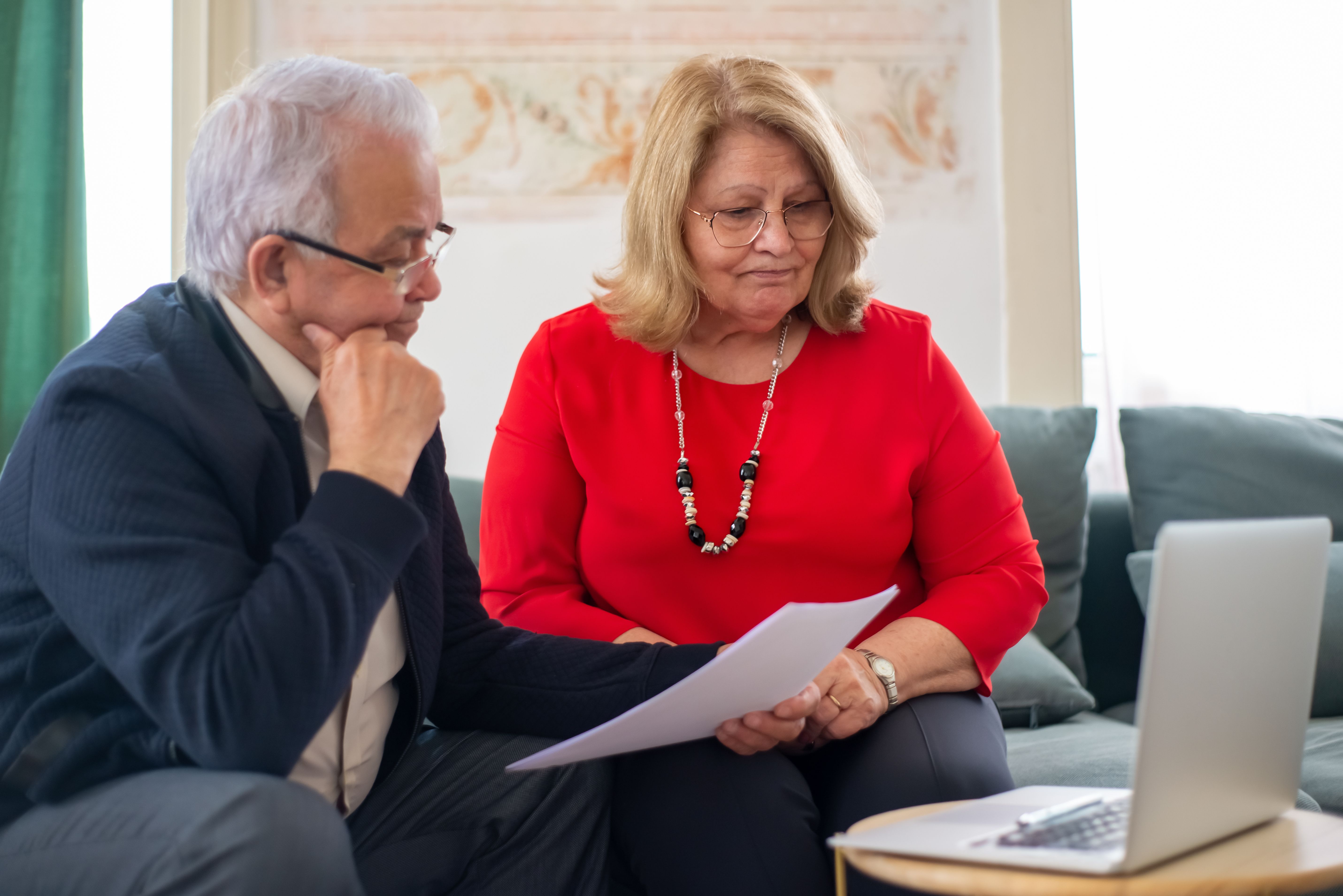 An elderly couple sitting on a sofa looking concerned while reviewing a printed document in front of a laptop.