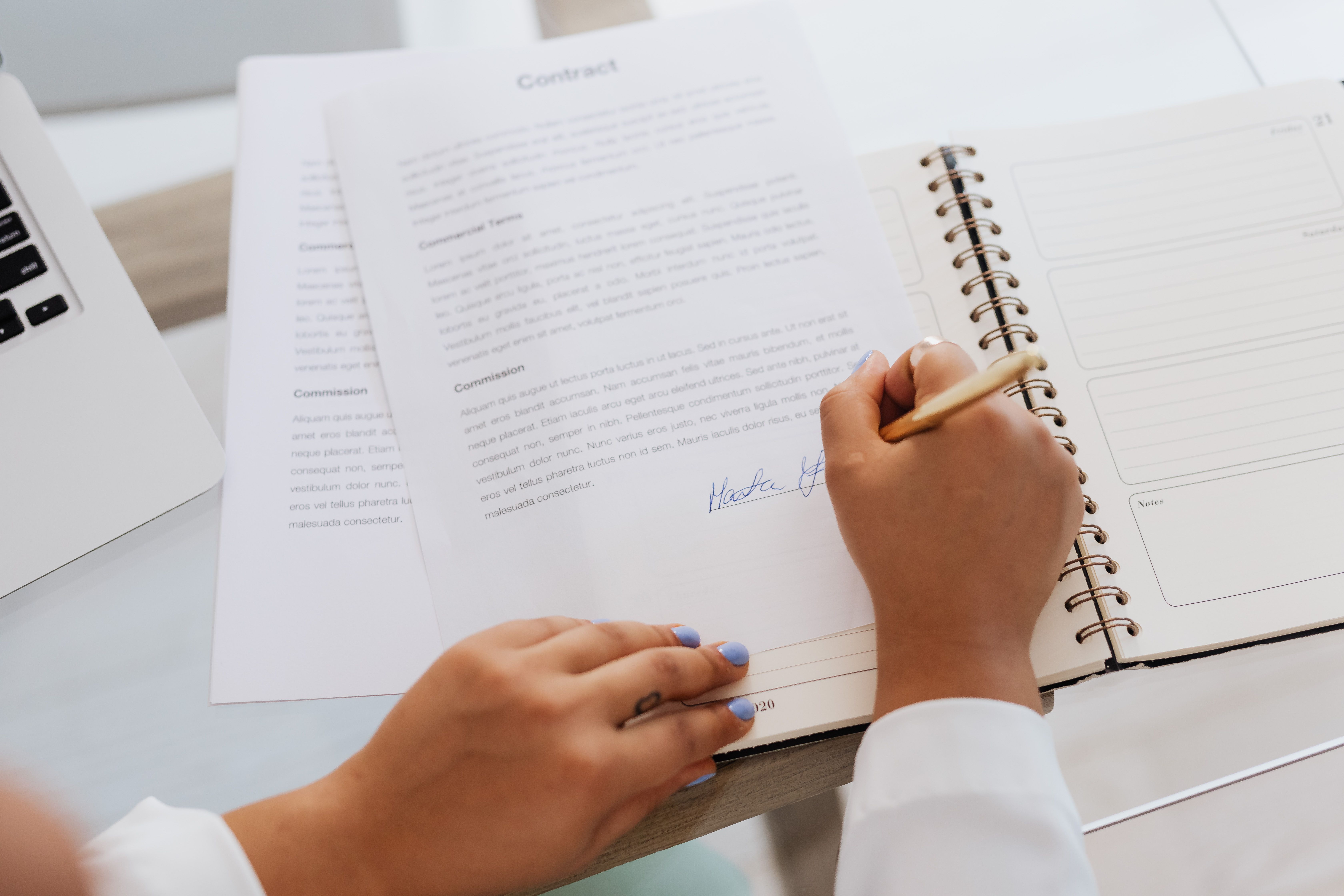 Close-up of a person holding a gold pen while signing a printed contract next to an open notebook.