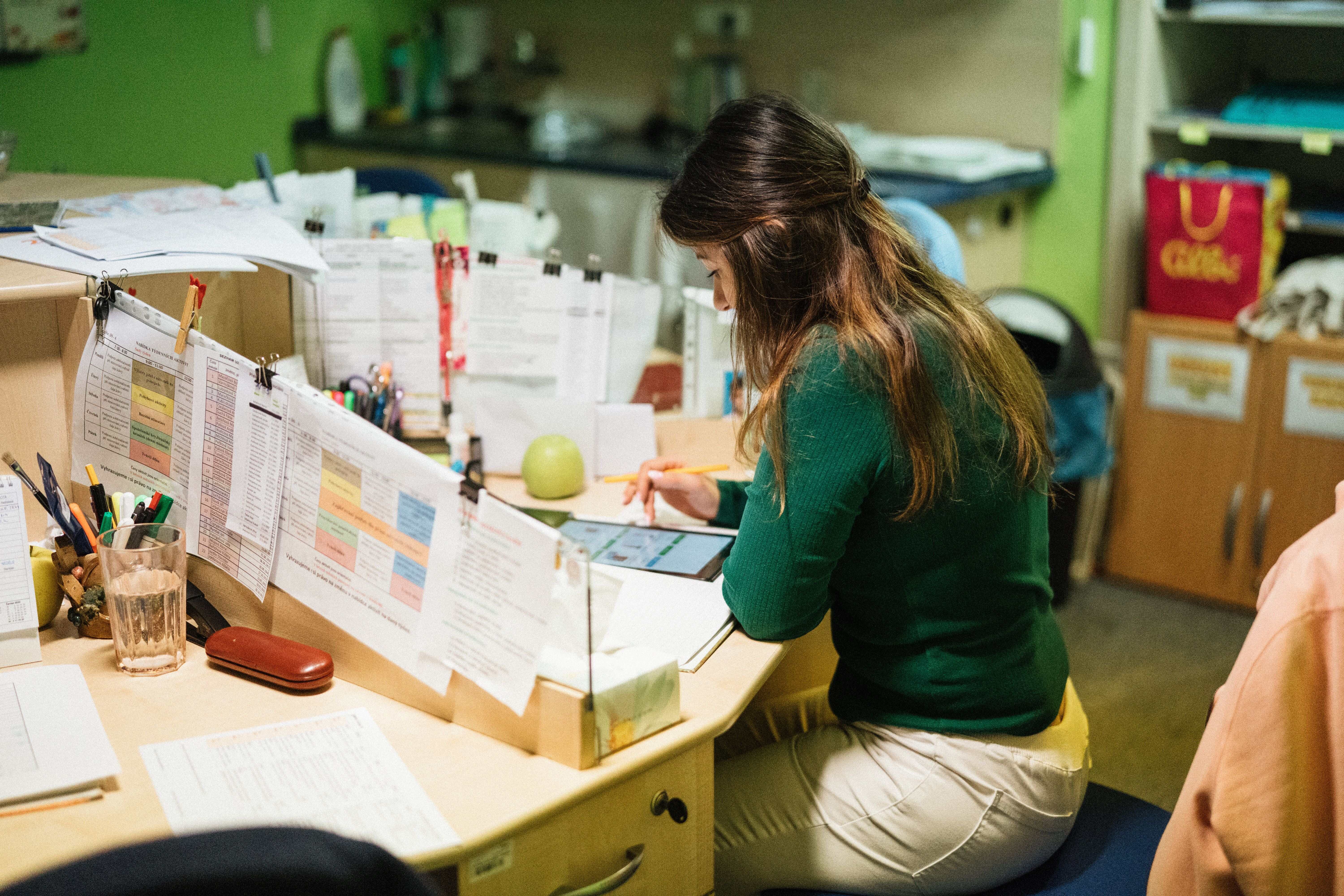 A woman sitting at a busy office desk filled with printed schedules, notes, and a tablet.