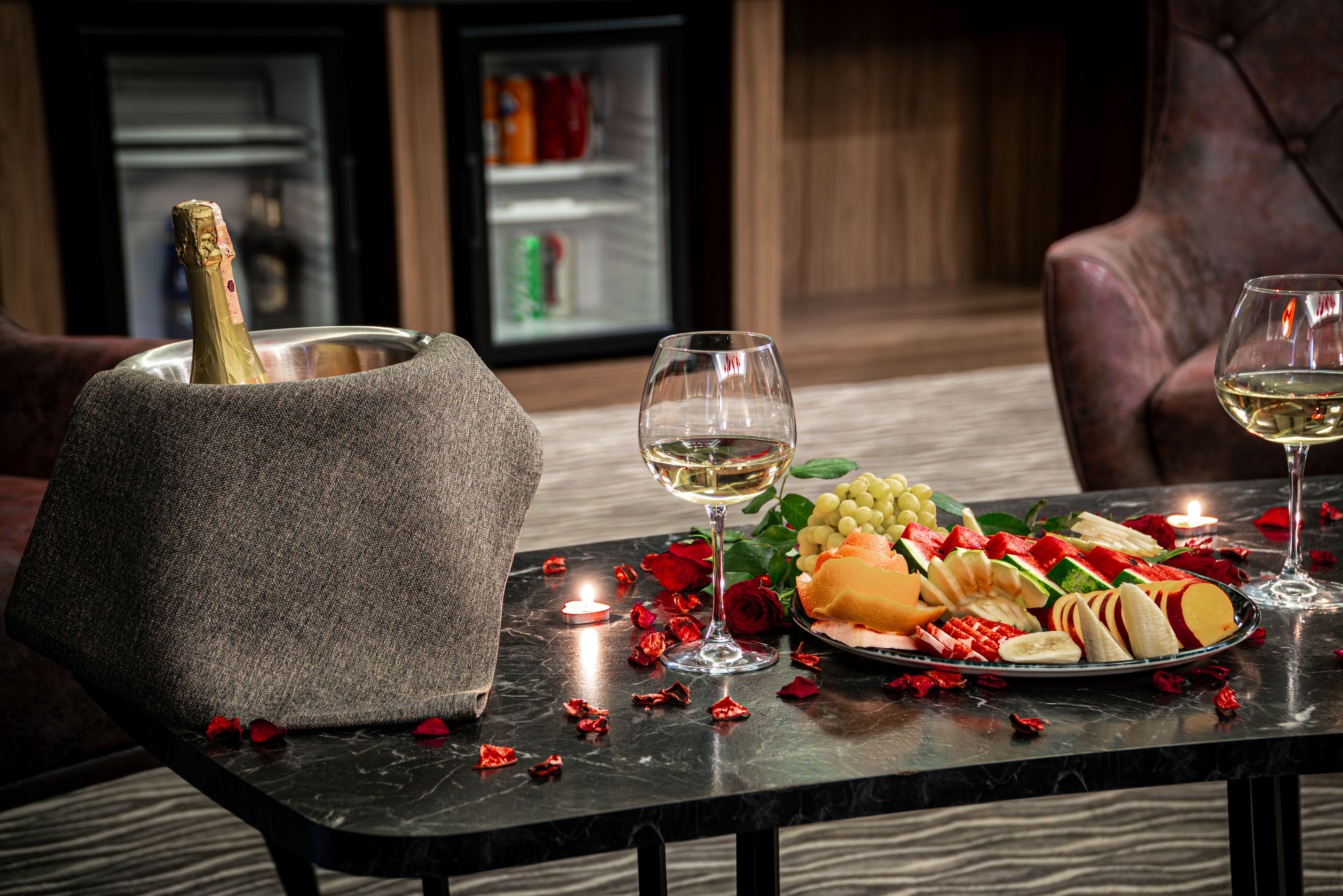 A tray of fresh sliced fruits, wine glasses, and a champagne bucket set on a table in front of mini refrigerators.