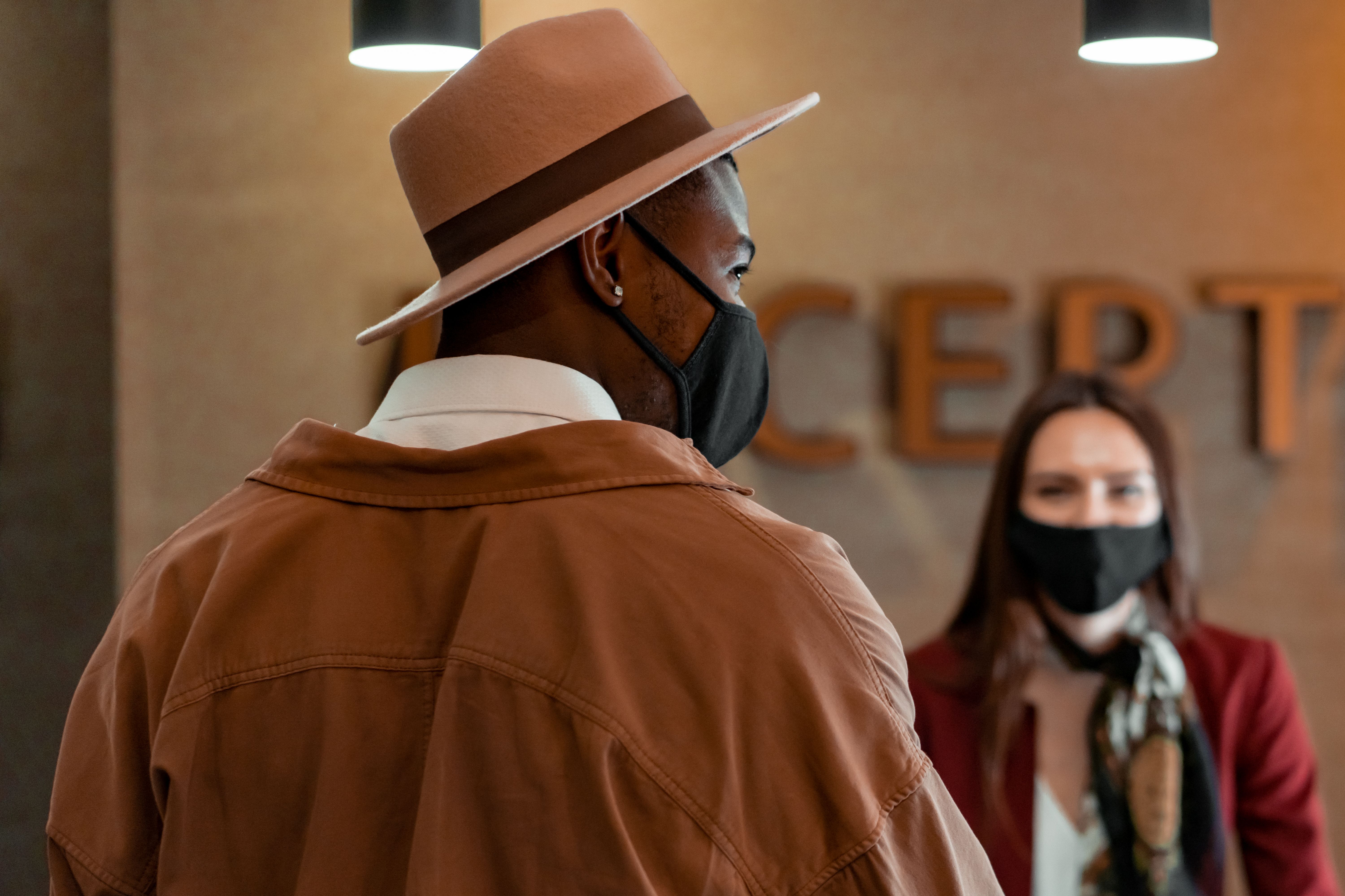 A man wearing a hat and black face mask speaking with a female receptionist in a hotel lobby.