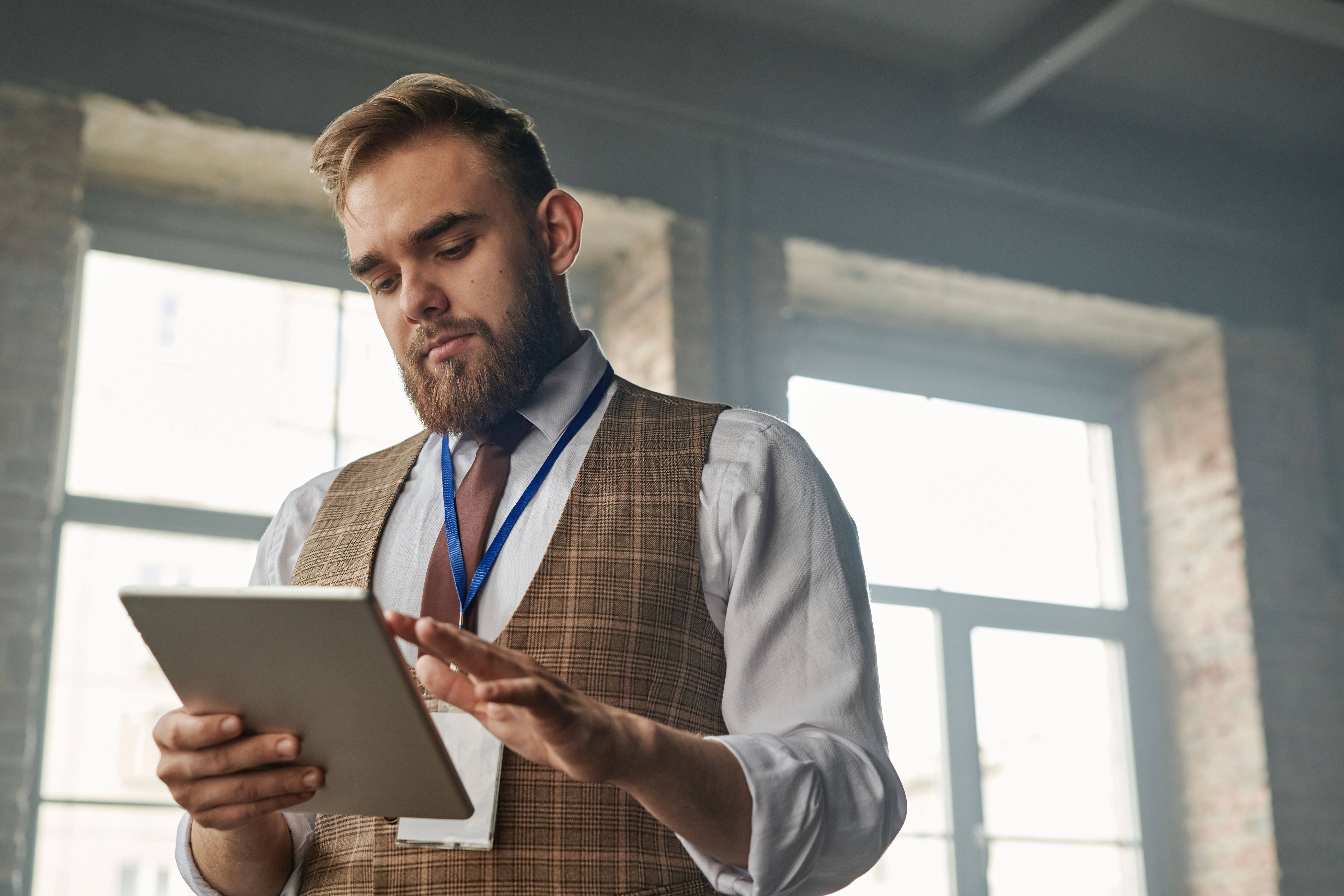 A man wearing a plaid vest, tie, and lanyard ID badge looking down while operating a digital tablet indoors.