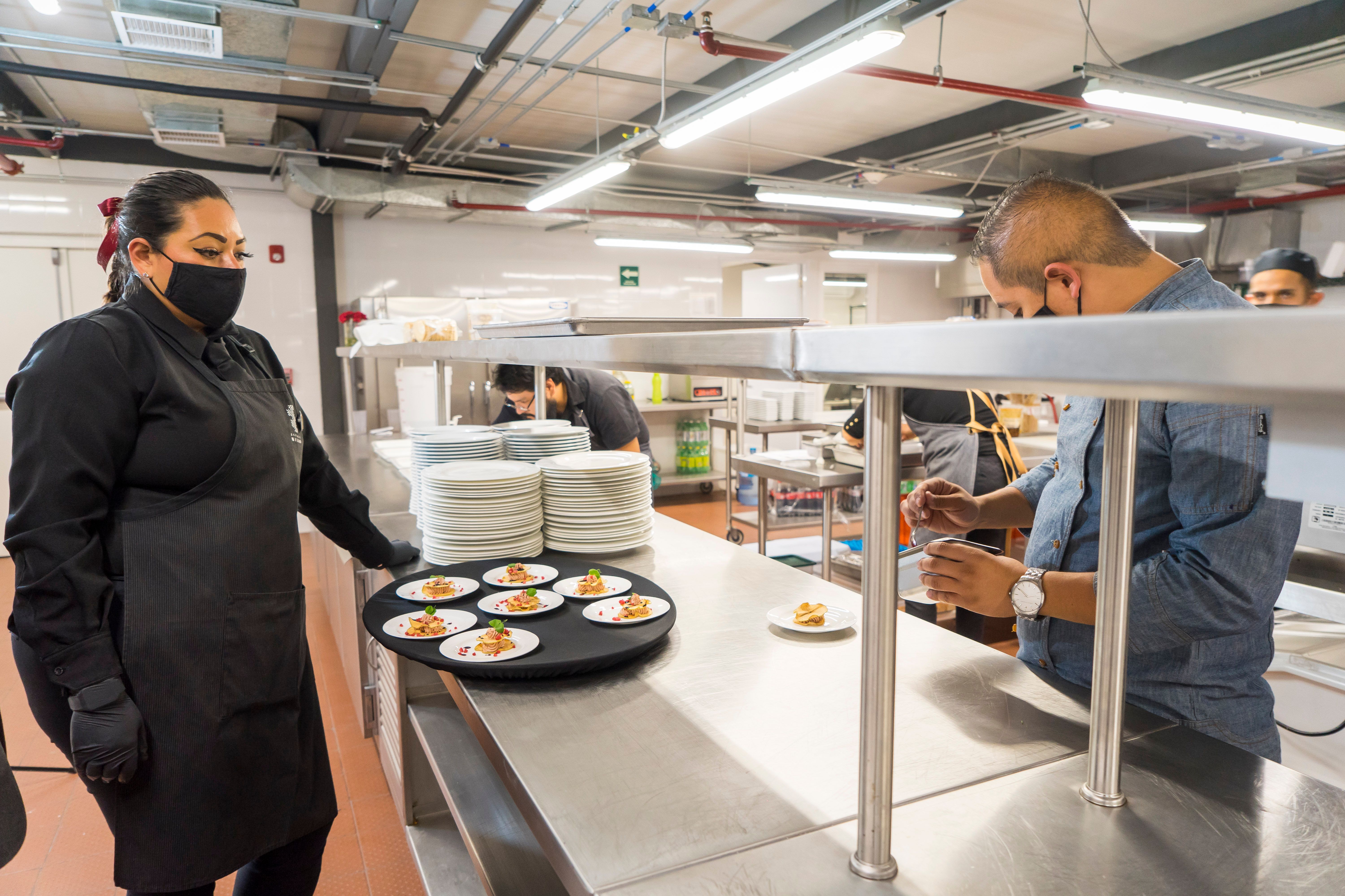 A kitchen staff member wearing a black mask and apron watching a chef prepare plated appetizers in a commercial kitchen.