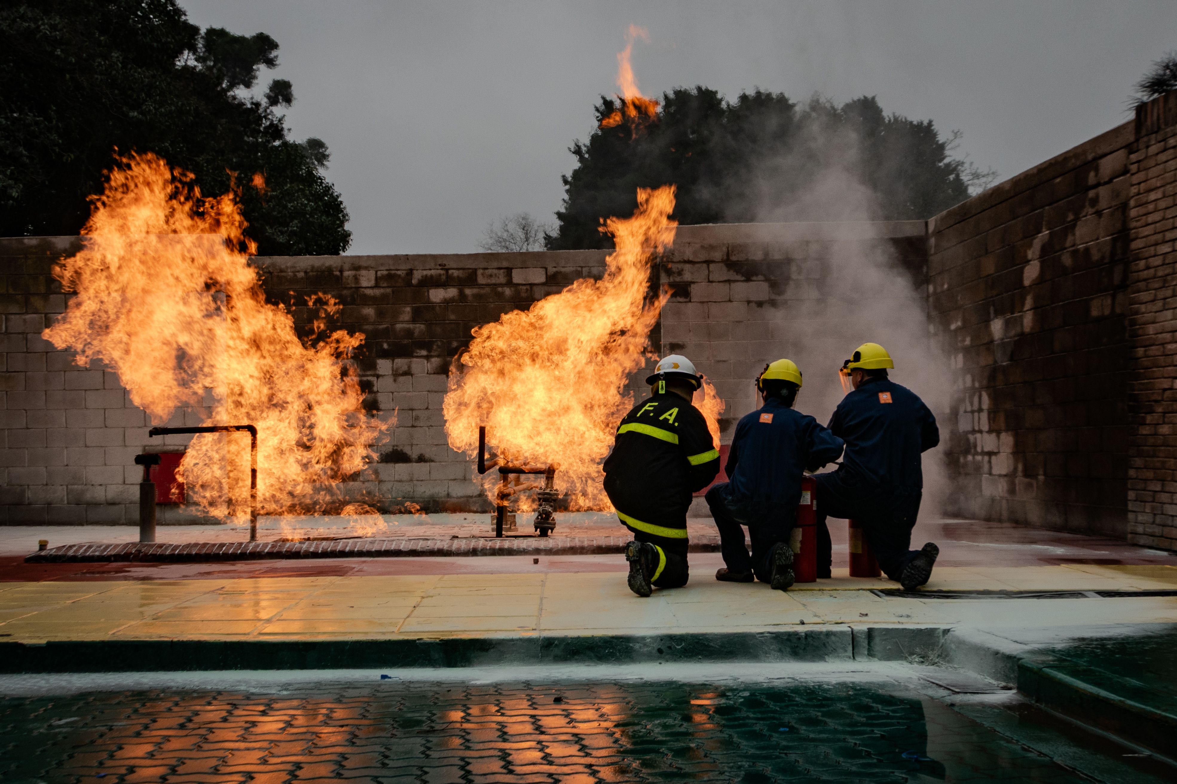 Three firefighters kneeling in safety gear and helmets during a controlled fire training exercise outdoors.