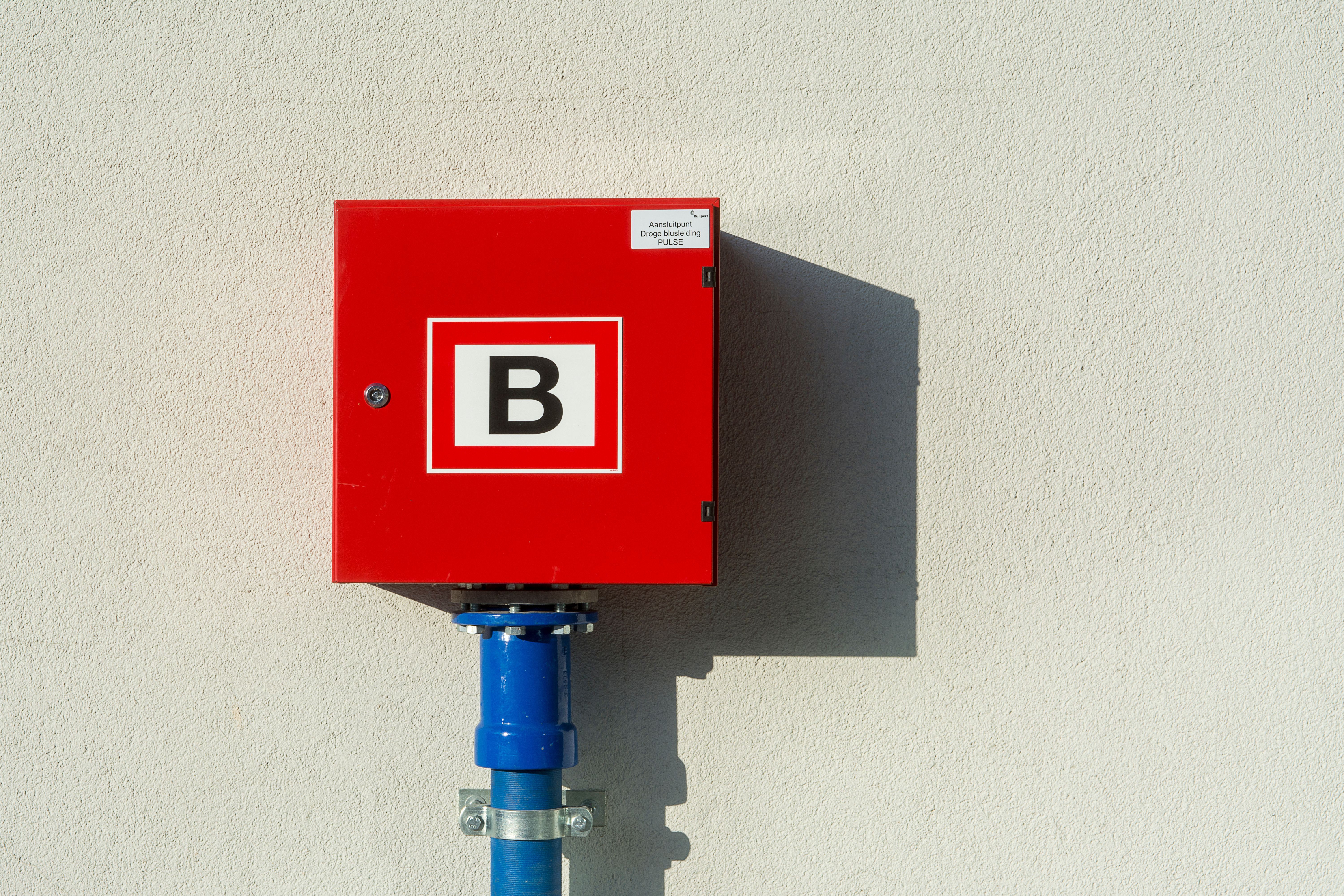 A red wall-mounted fire safety connection box labeled with a large letter B attached to a blue pipe.