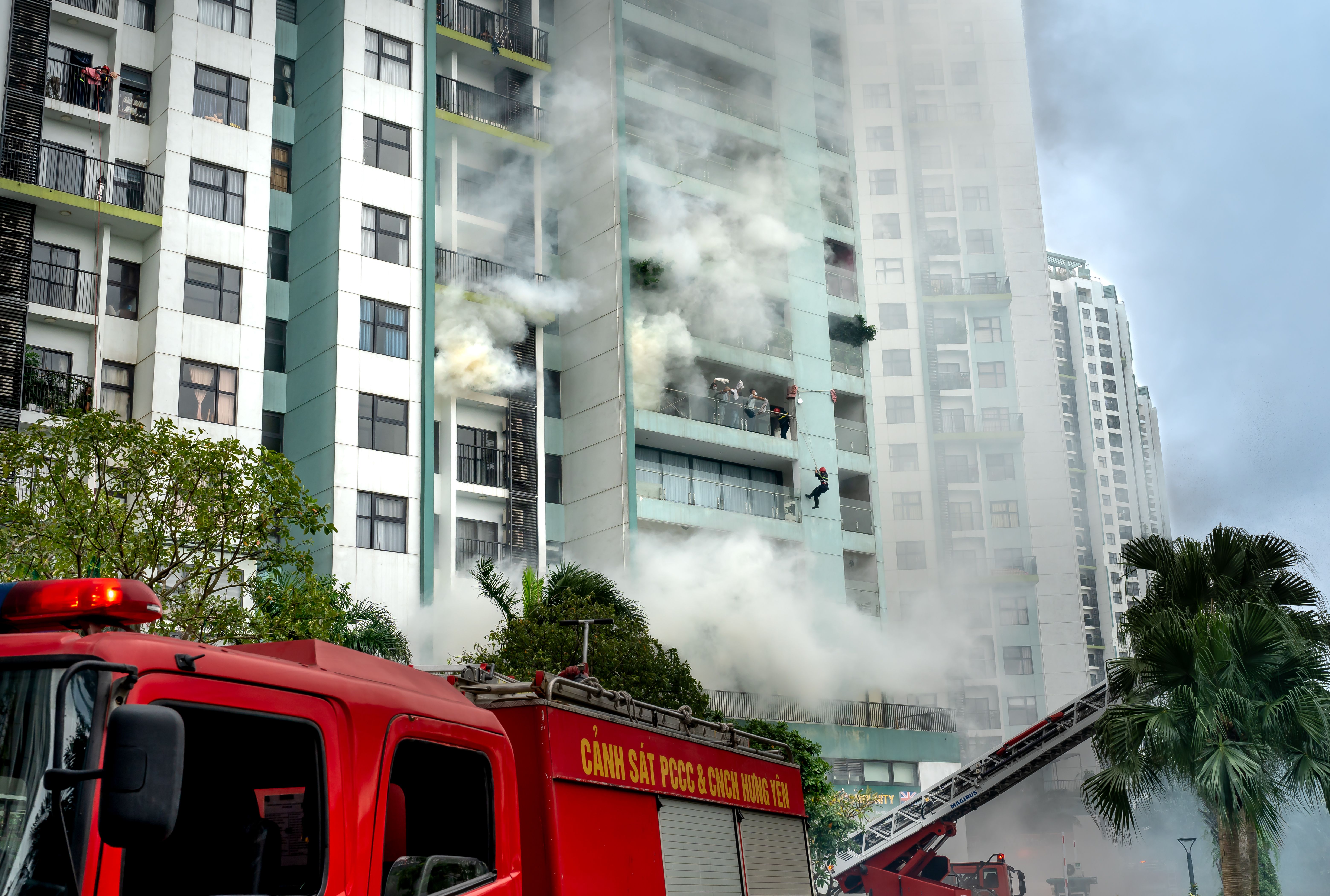Smoke rising from a multi-story residential building while firefighters conduct a balcony rescue with a ladder truck in the foreground.