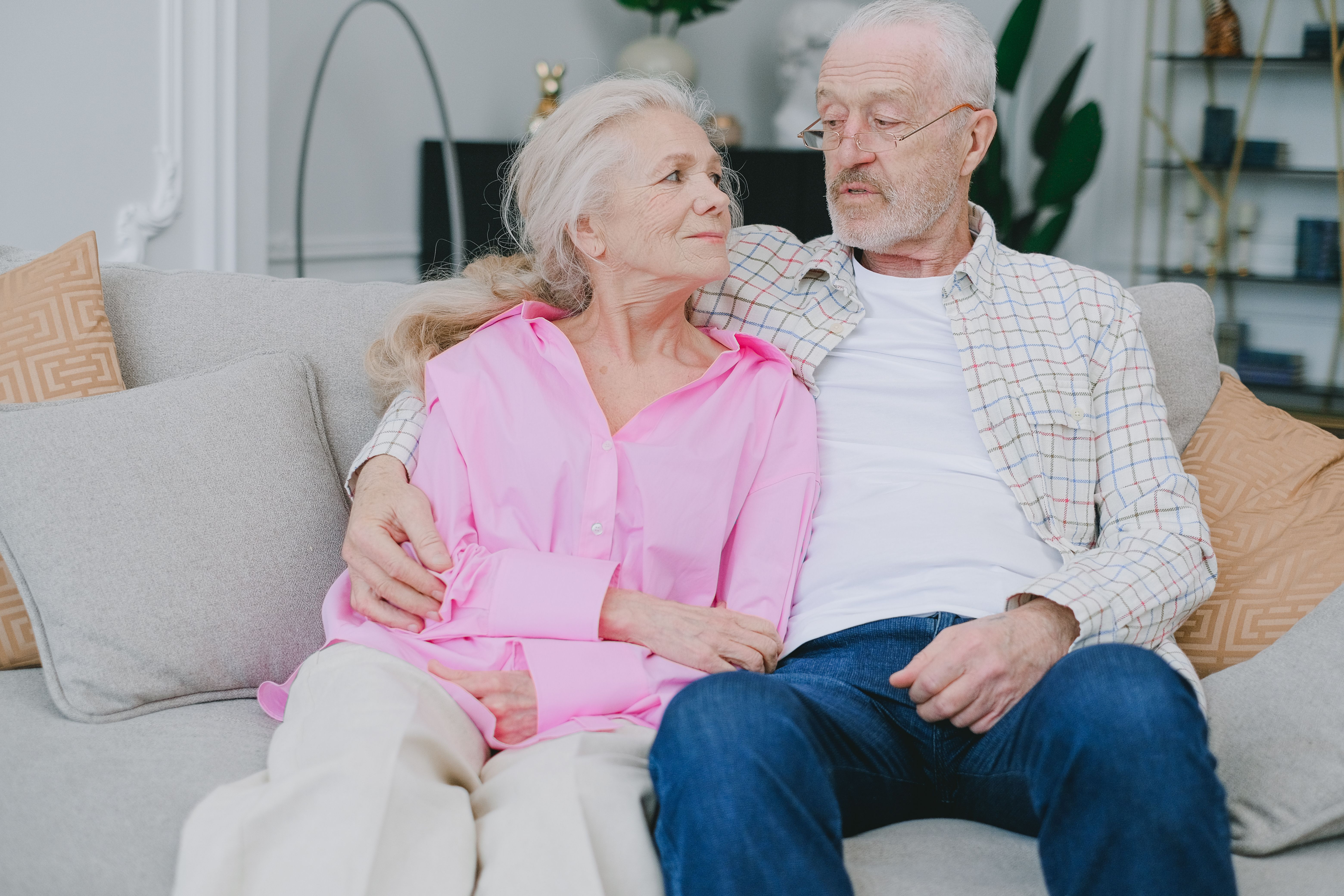 An elderly couple sitting closely together on a neutral fabric sofa in a bright living area.