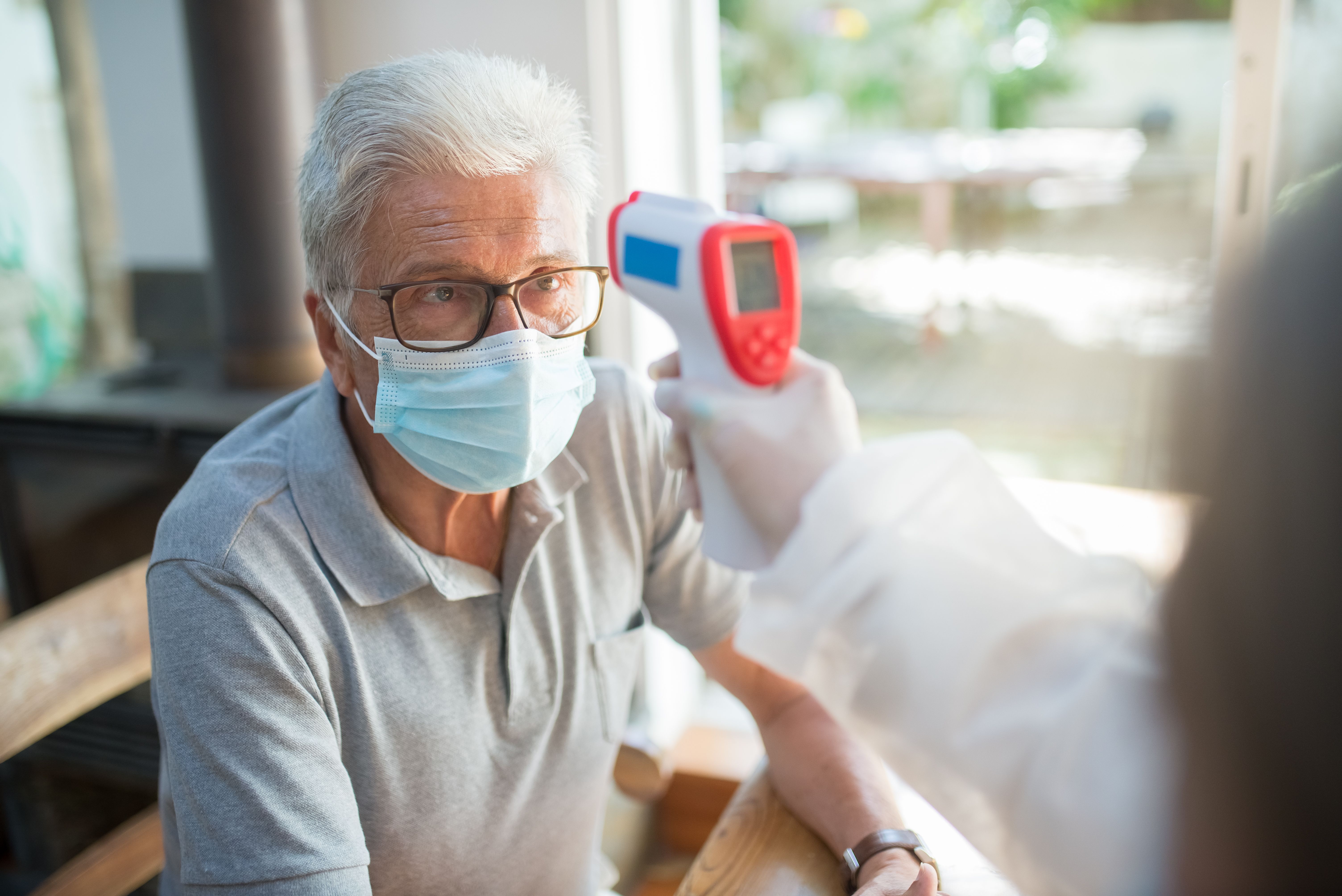 A healthcare worker in a white gown and gloves holding a digital infrared forehead thermometer to check the temperature of an older man wearing a face mask and glasses.