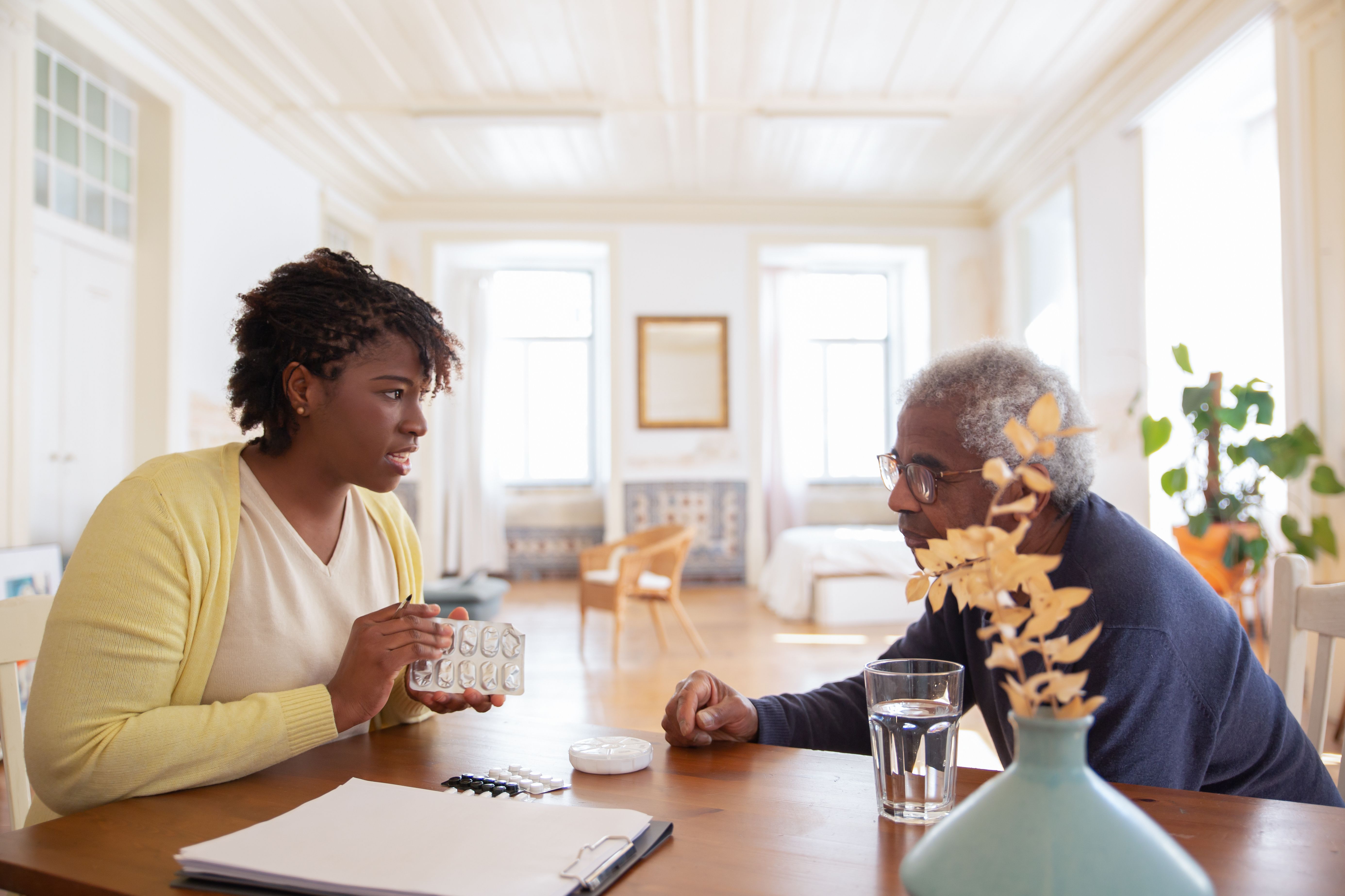 A female caregiver sitting at a table discussing medication blister packs with an elderly man.