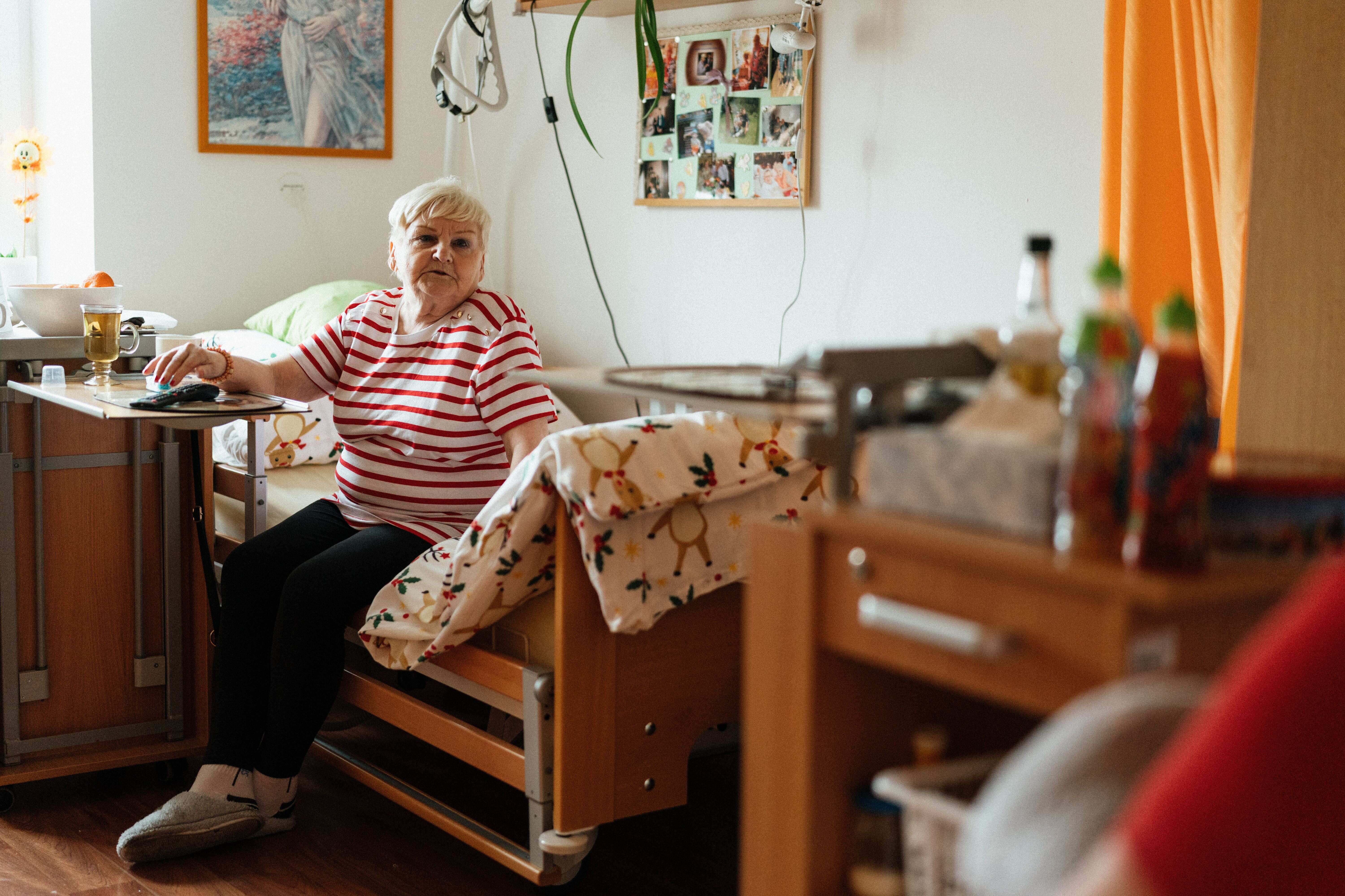 An elderly woman in a red-and-white striped shirt sitting on the edge of an adjustable bed inside a care home room.