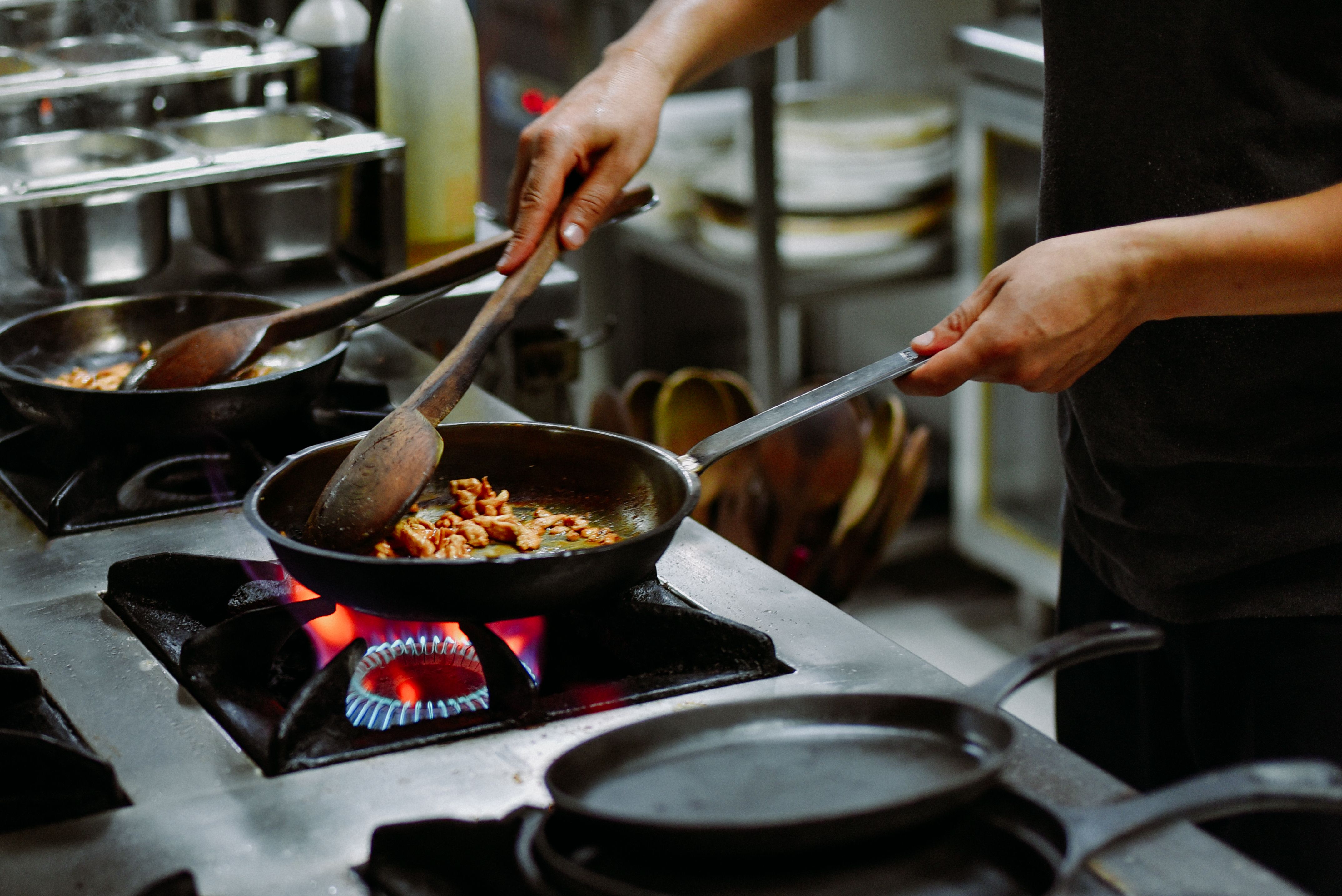 A chef holding a wooden spoon while stir-frying food in a pan over a gas flame in a commercial kitchen.