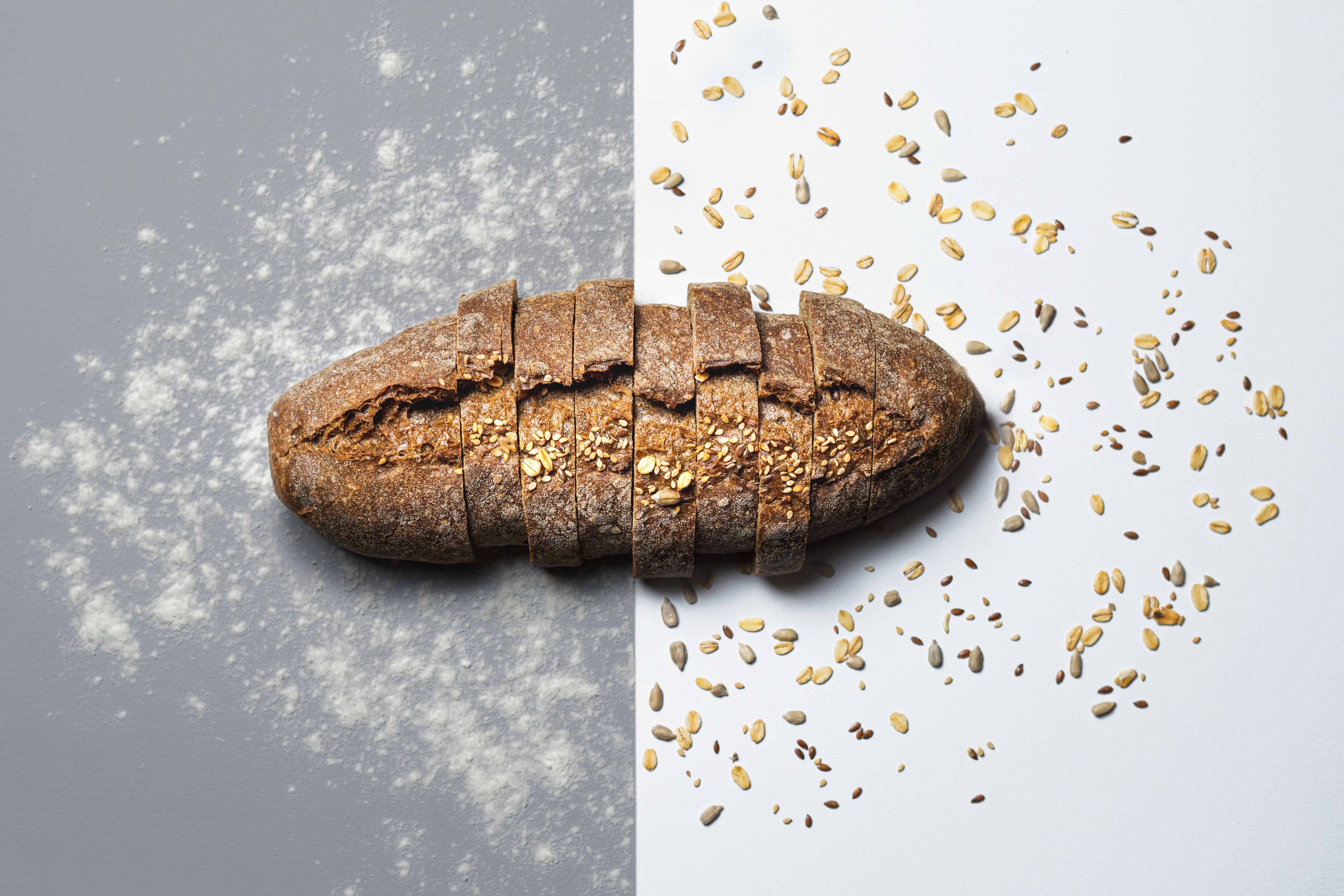 A sliced whole-grain bread loaf centered on a split background with flour dust on the left and scattered seeds on the right.