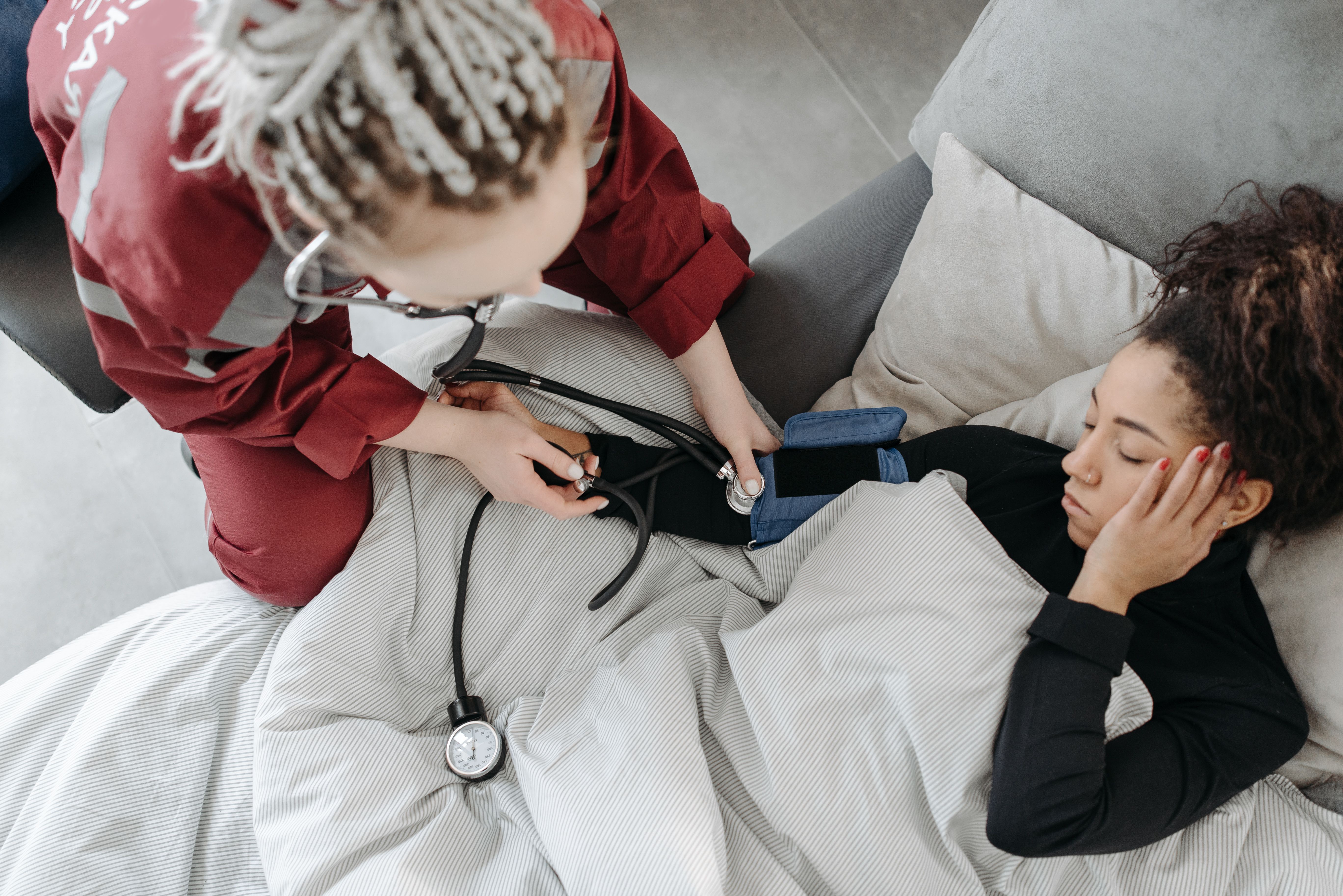 A medical responder in red uniforms measuring the blood pressure of a woman resting on a sofa under a blanket.