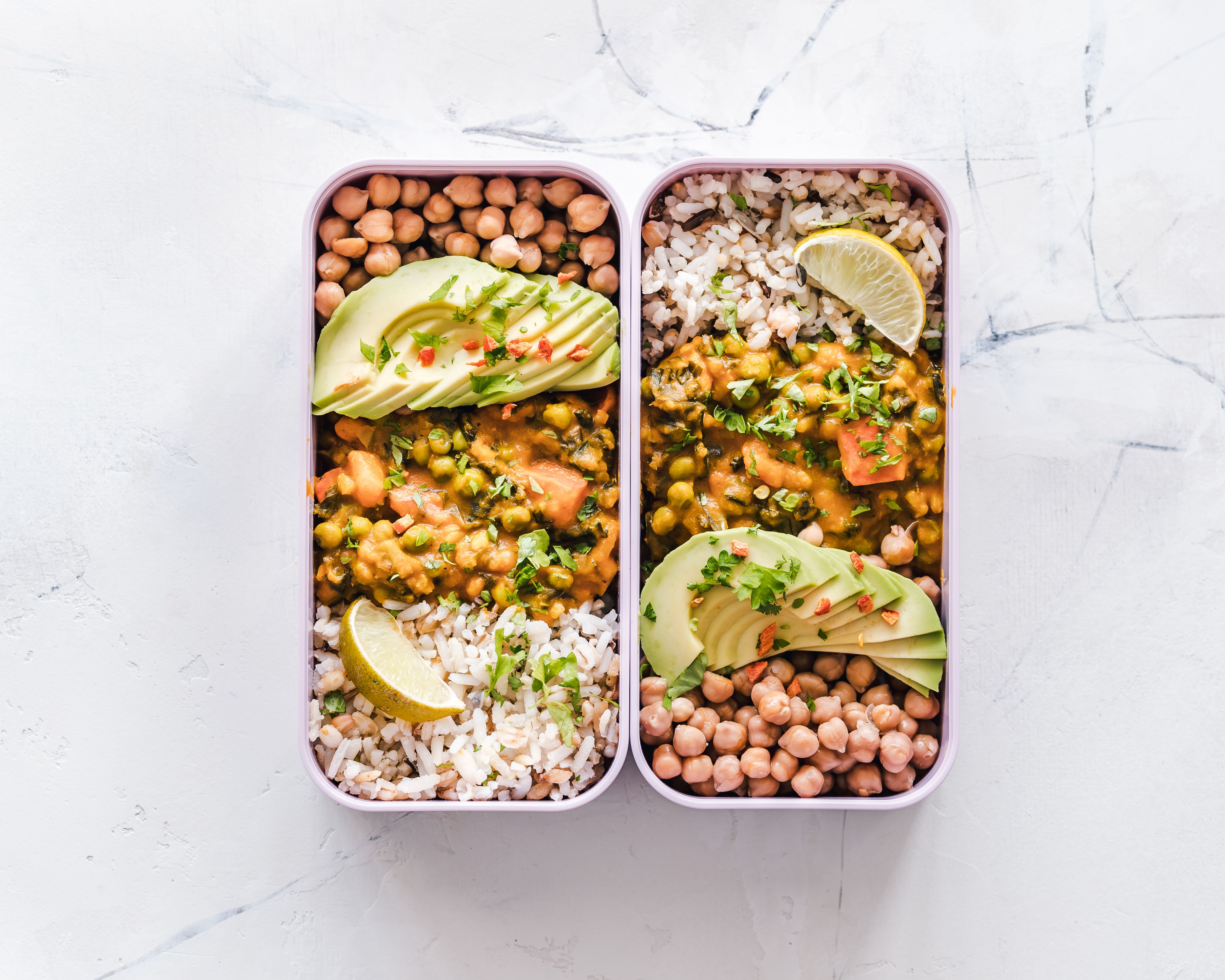 Two prepped meal containers filled with rice, chickpea curry, sliced avocado, and fresh lime wedges.