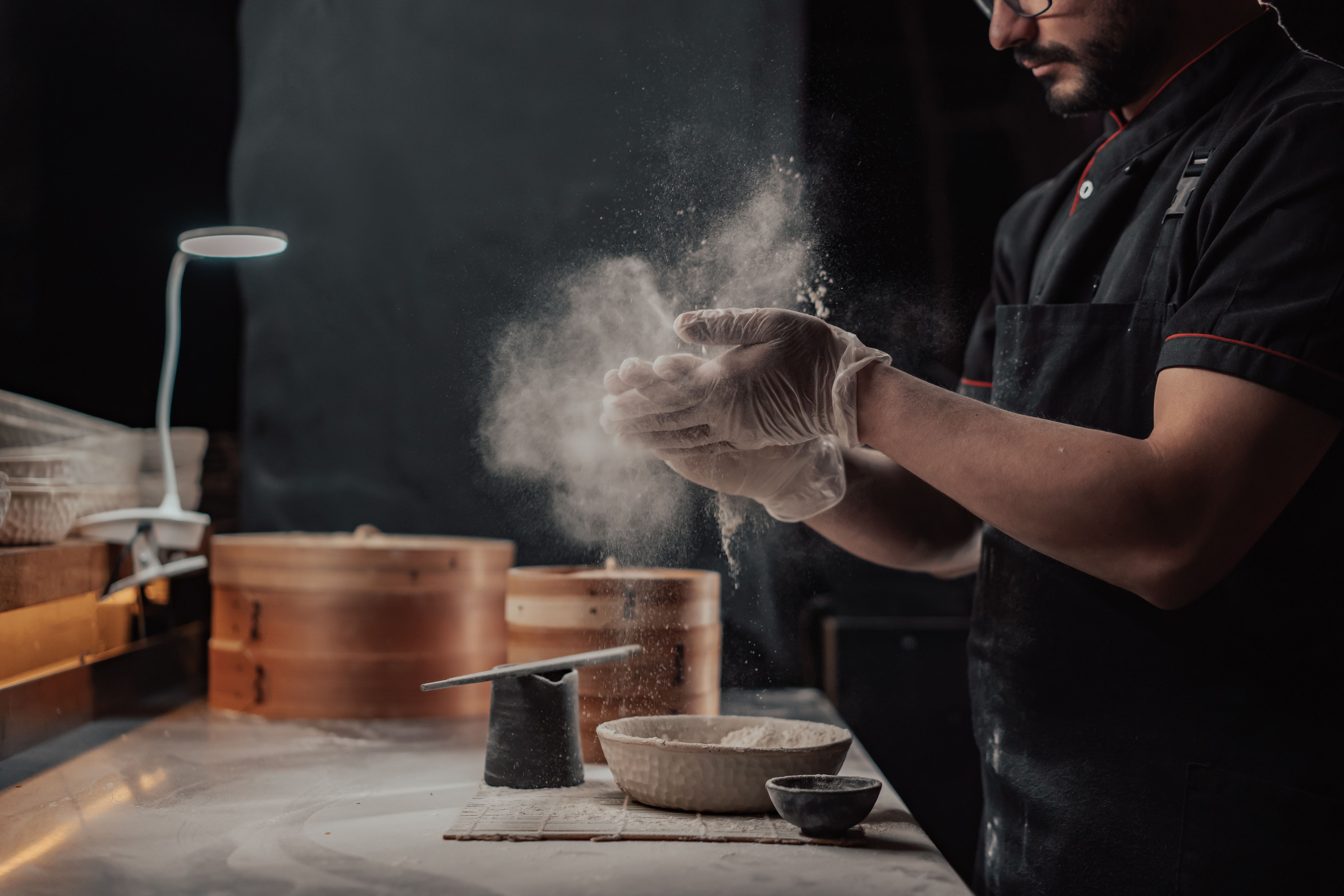 A chef wearing gloves clapping flour off their hands in a commercial kitchen workspace.