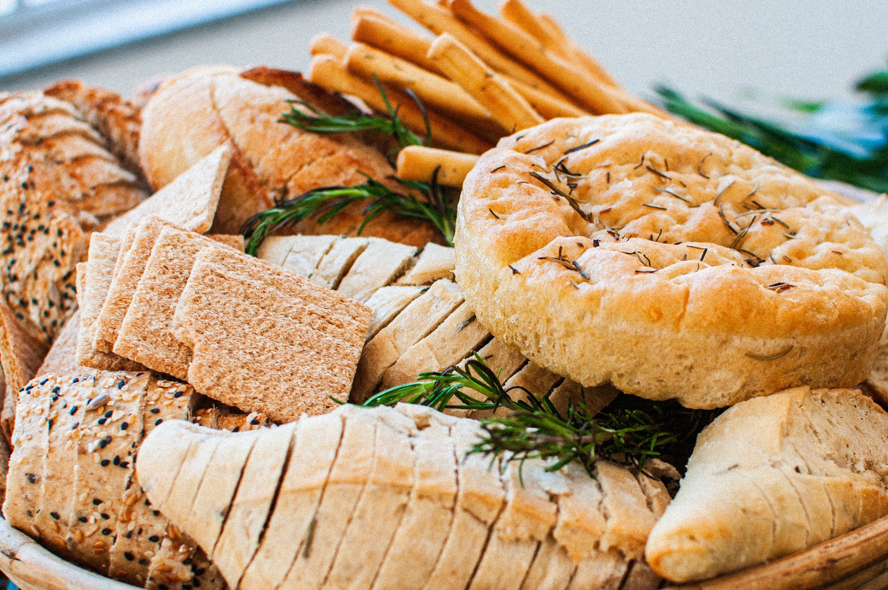 A bread platter featuring sliced loaves, rosemary focaccia, crackers, and breadsticks garnished with fresh rosemary sprigs.
