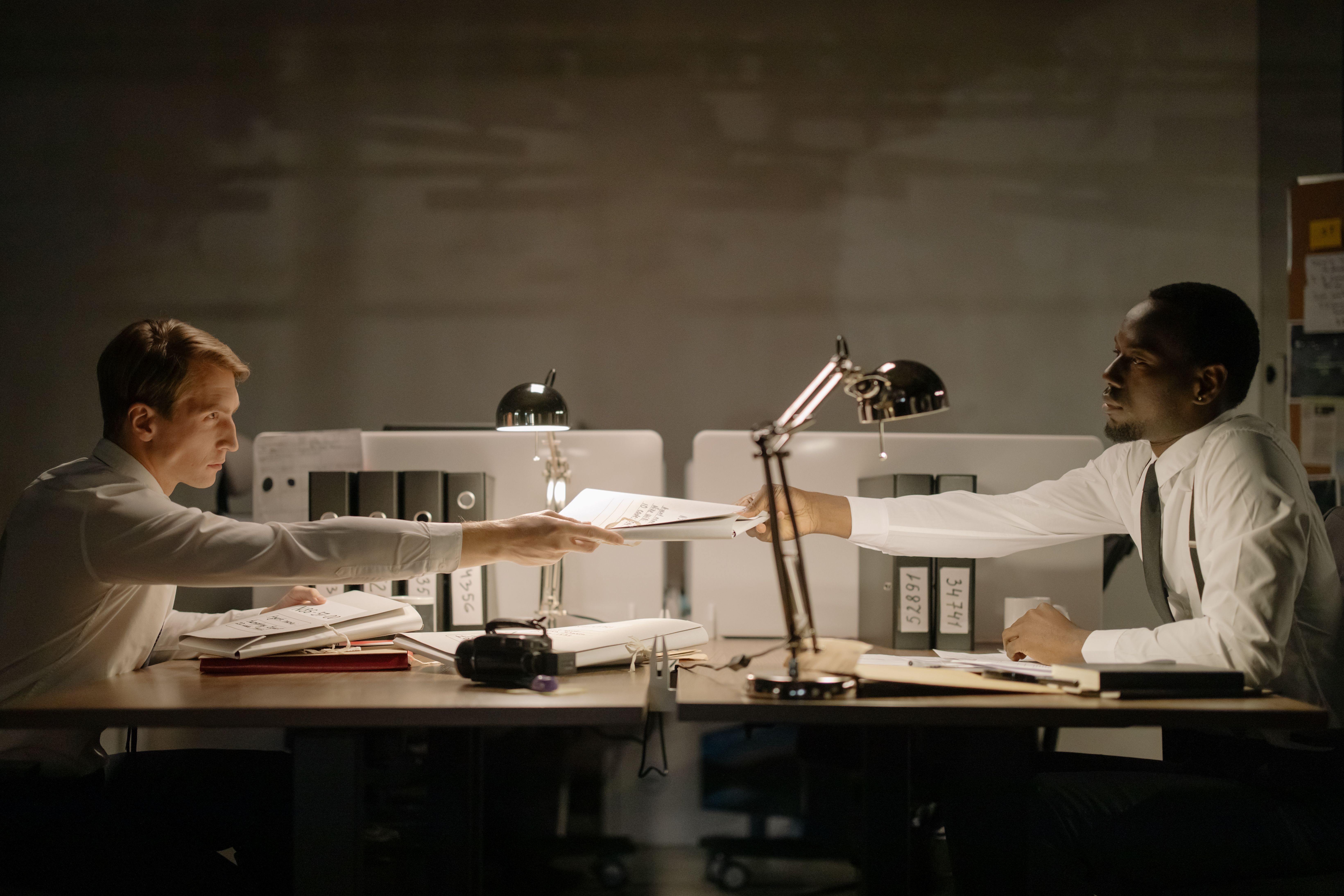Two office workers sitting across desks late at night exchanging a stack of paper files.