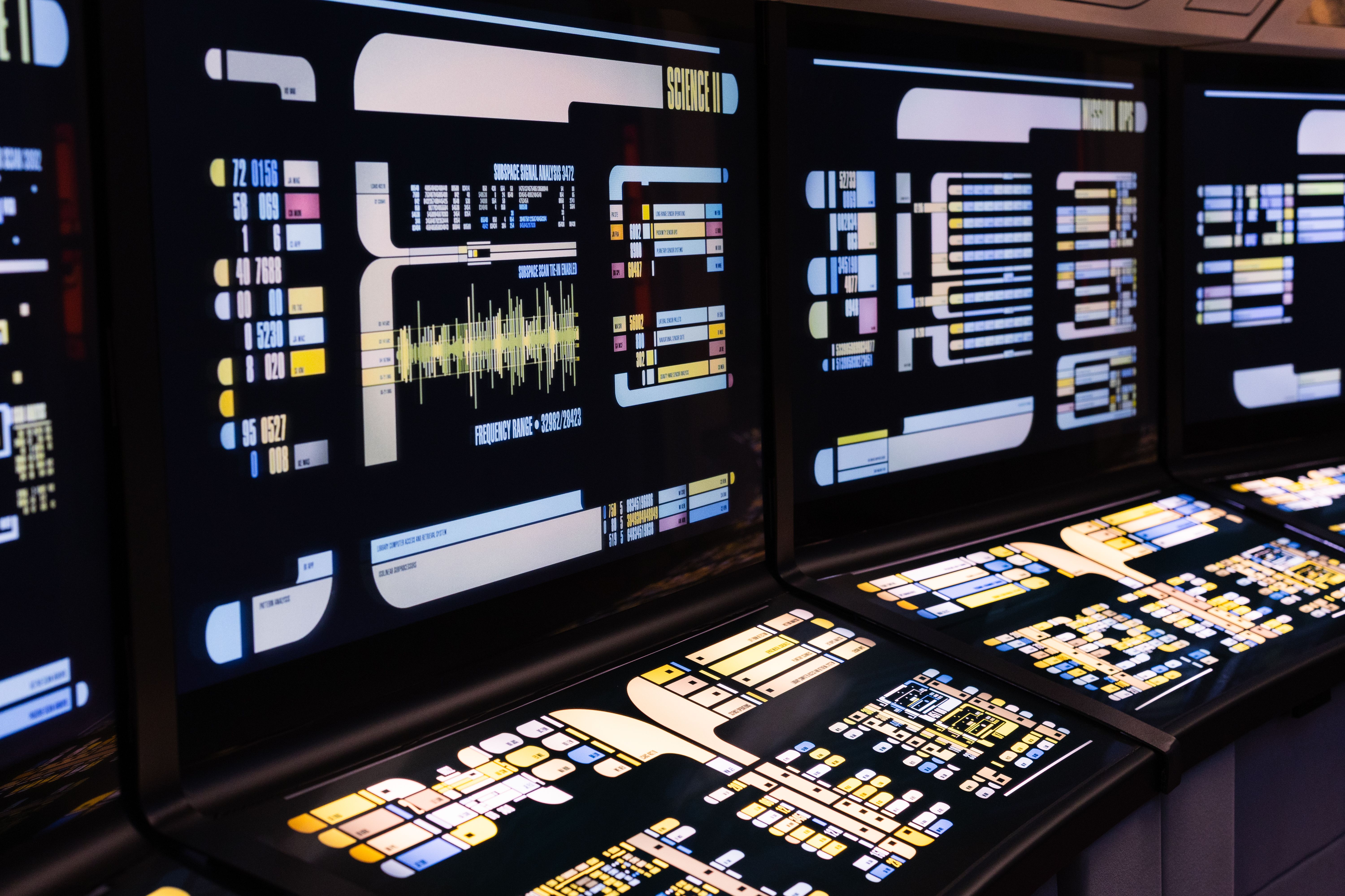 High-tech control room monitors displaying illuminated futuristic graphs, frequency waves, and system interface data.