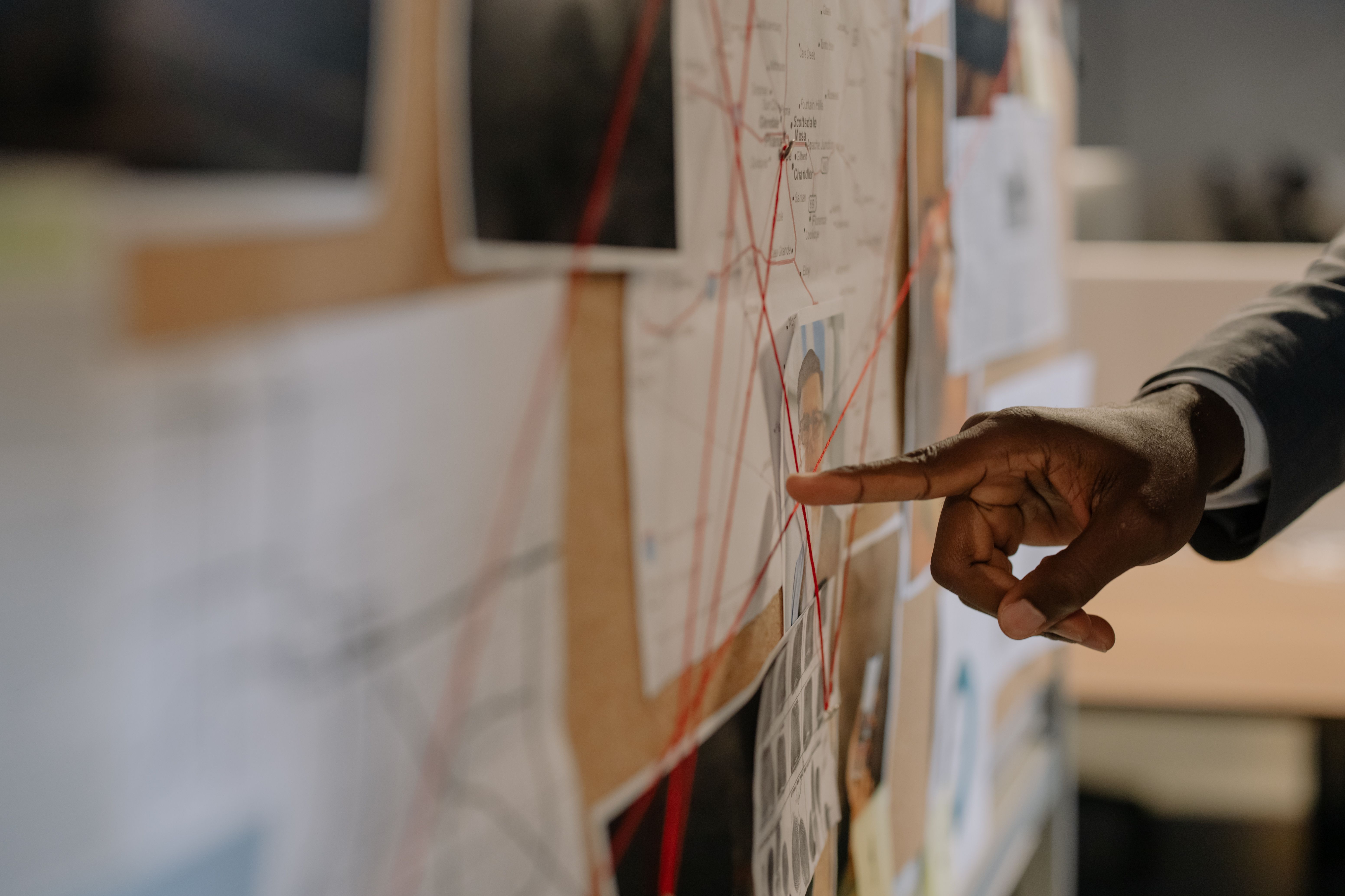 A close-up of a hand pointing to a photo pinned on an investigation board connected by red string.