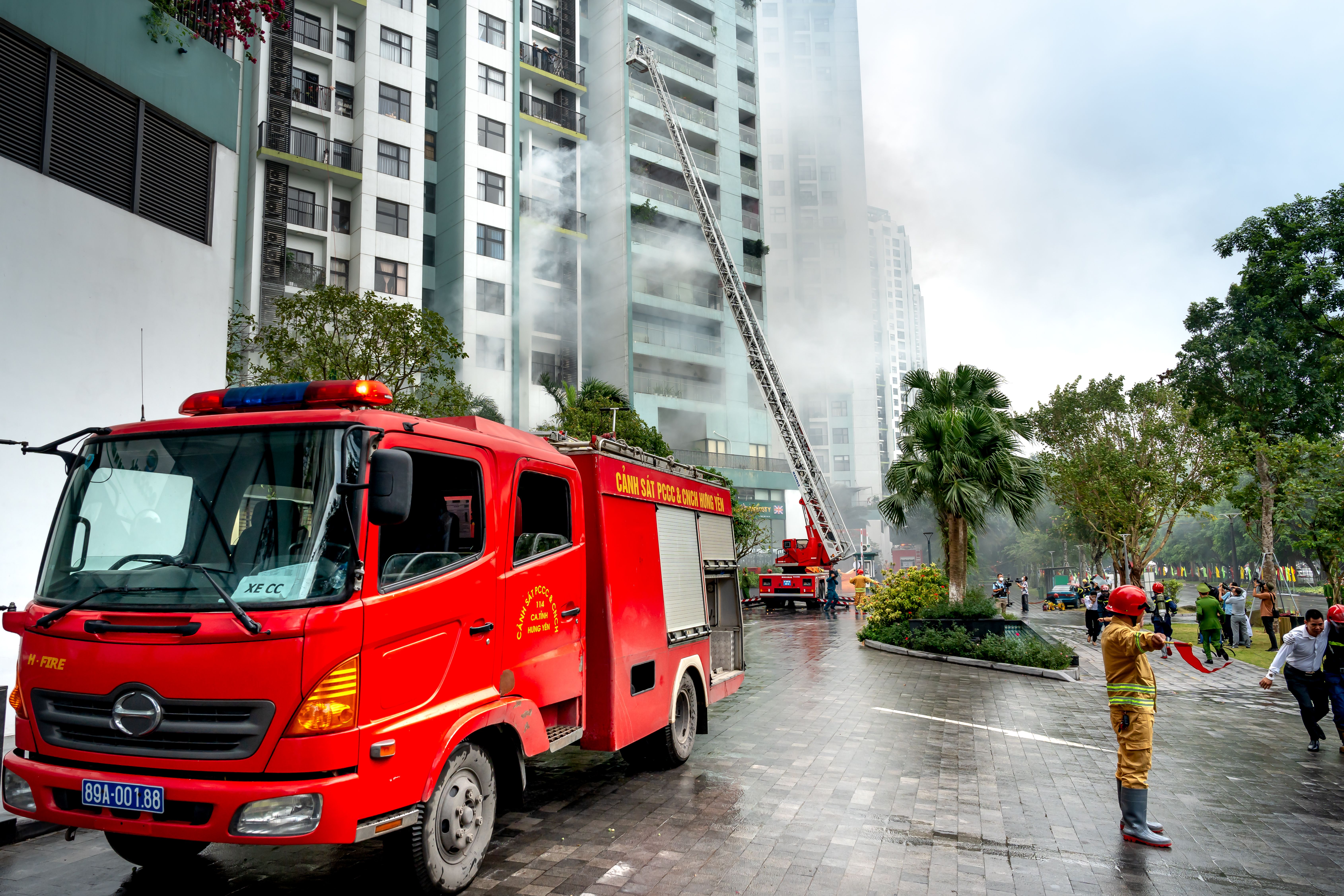A red fire engine parked outside a high-rise residential building with a ladder extended toward a smoking upper floor.