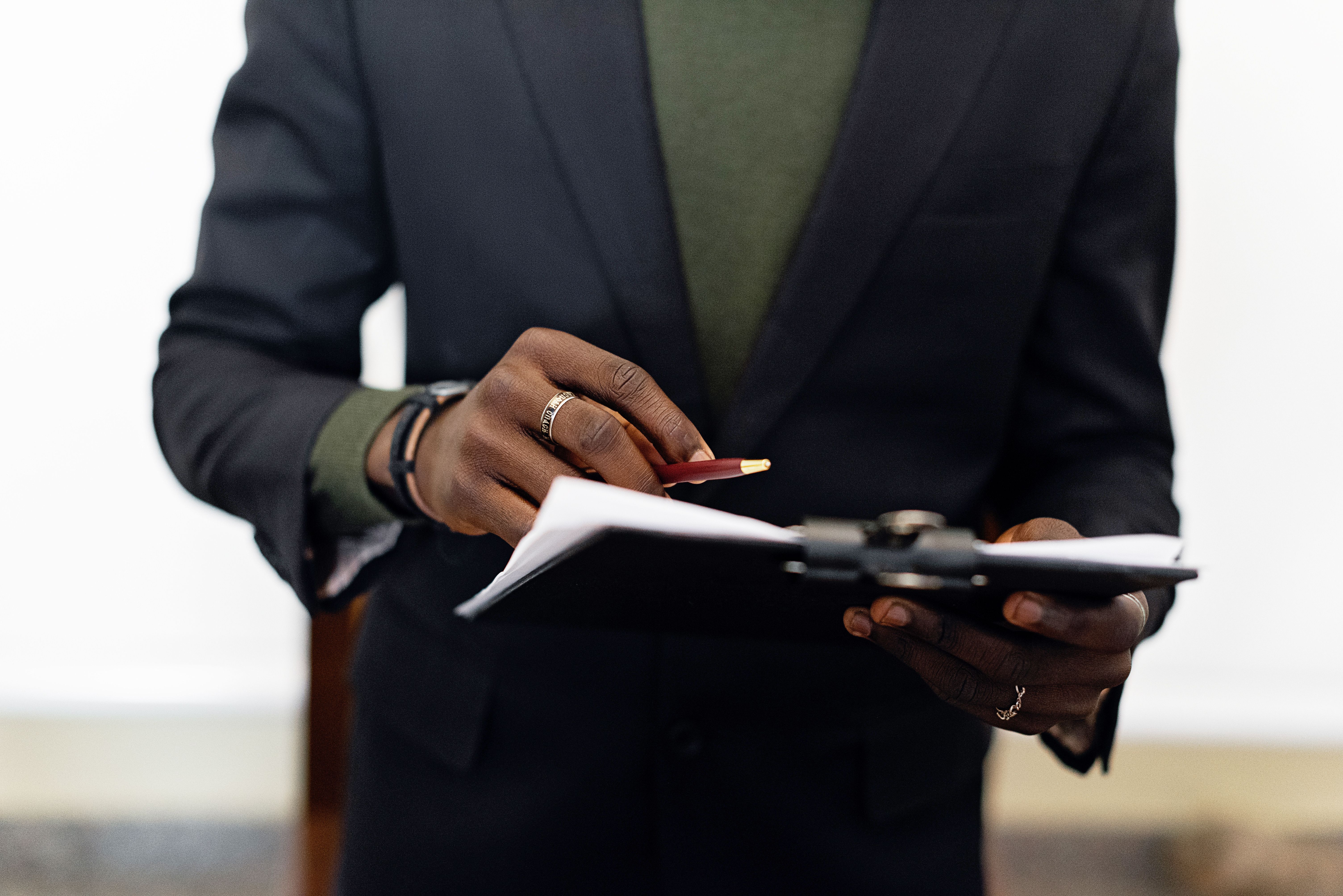 A man in a dark suit jacket holding a pen and reviewing documents on a clipboard.