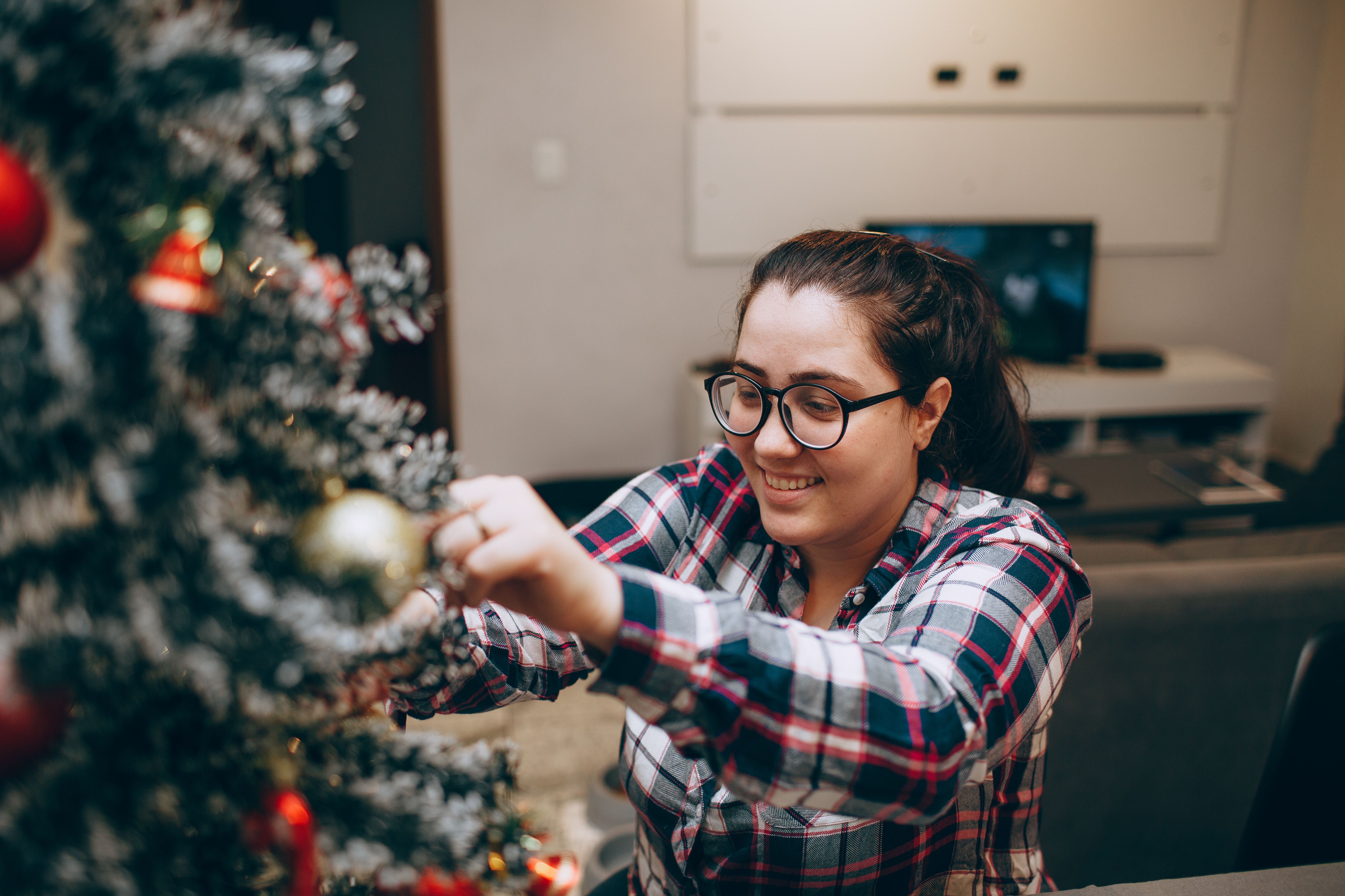 A woman in a plaid shirt hanging a golden bauble on a frosted Christmas tree indoors.