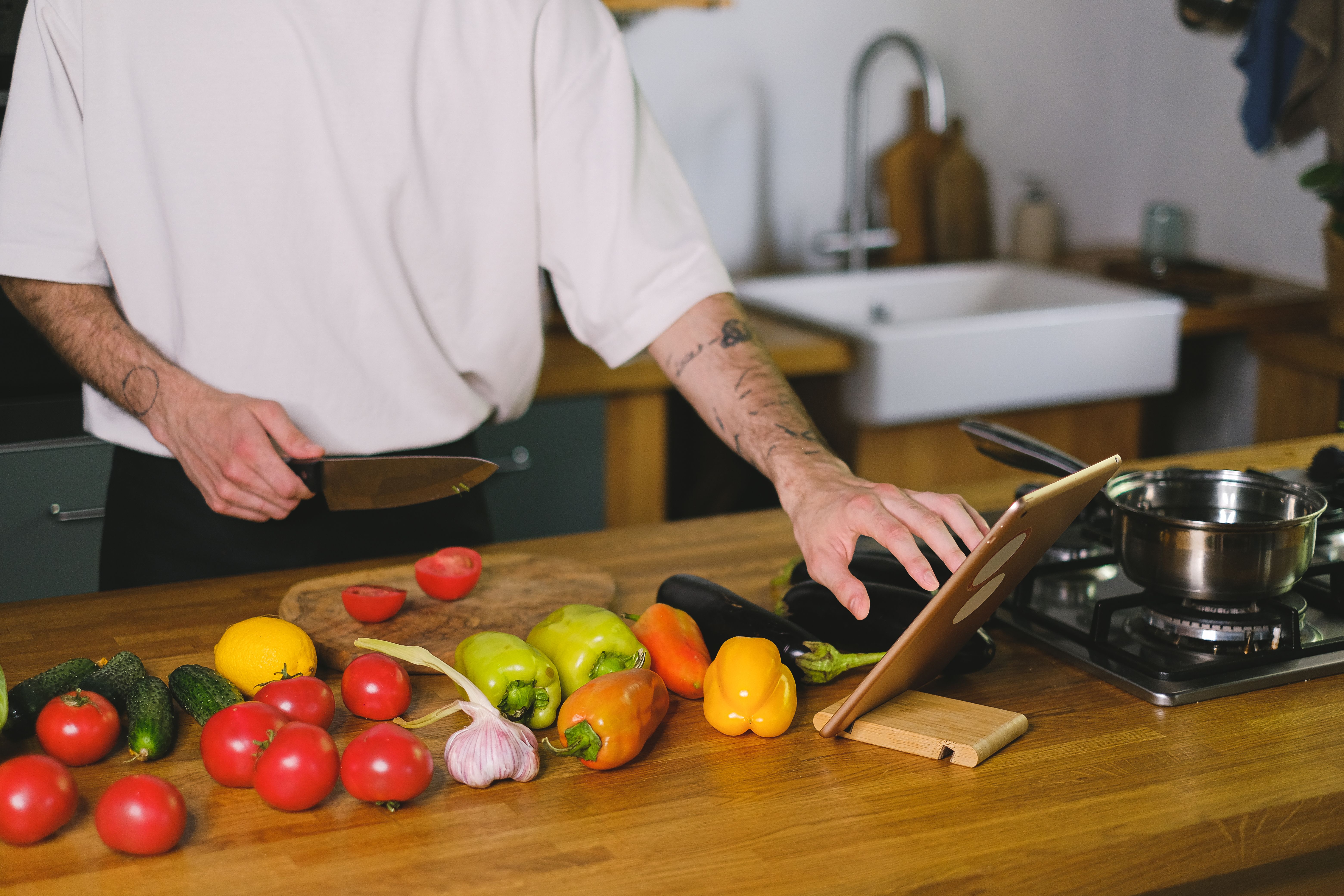 A person holding a chef's knife while using a tablet placed on a kitchen counter near fresh vegetables.