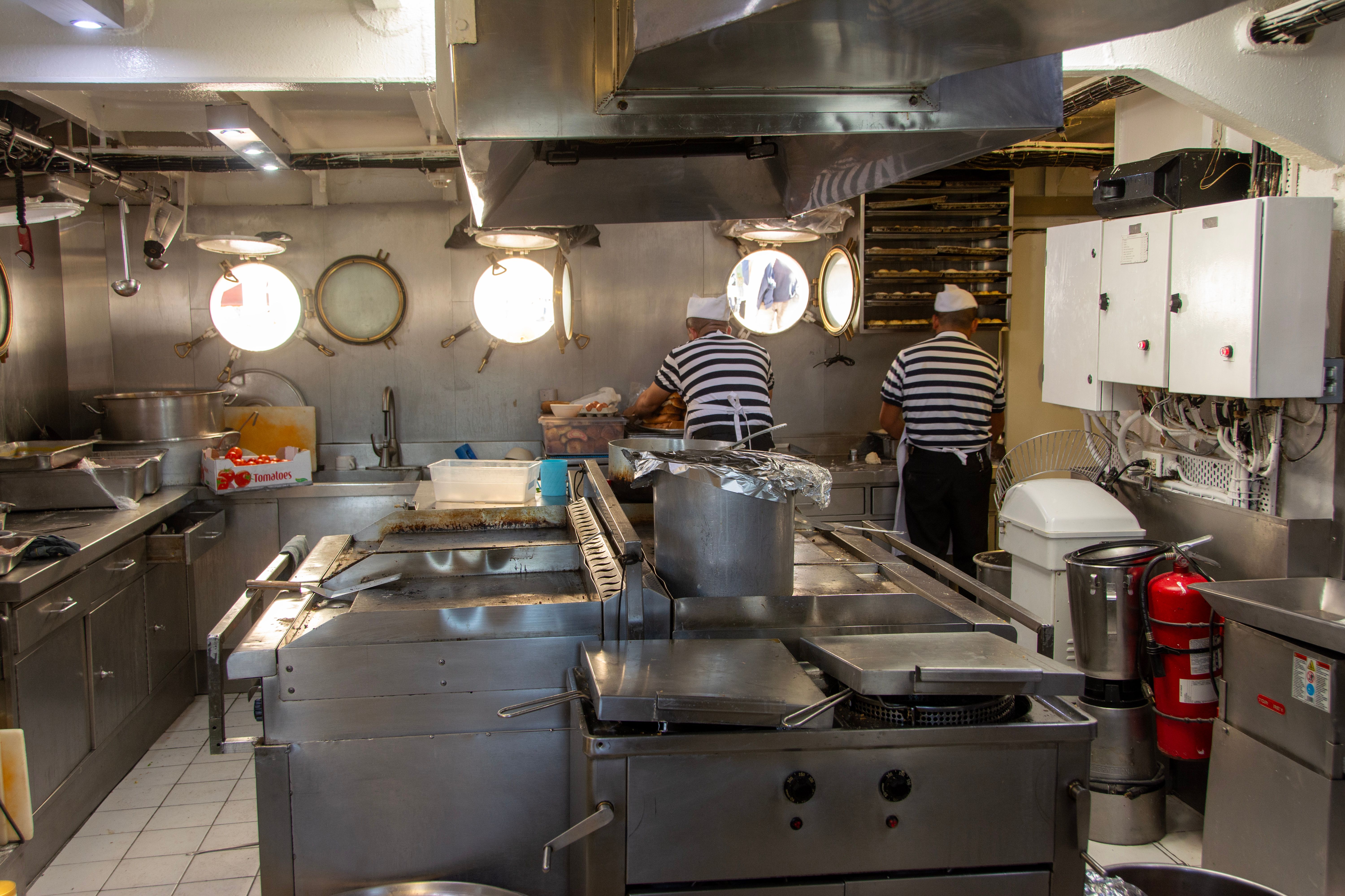 Two kitchen staff members preparing food inside a commercial stainless steel ship galley equipped with cooking stations.