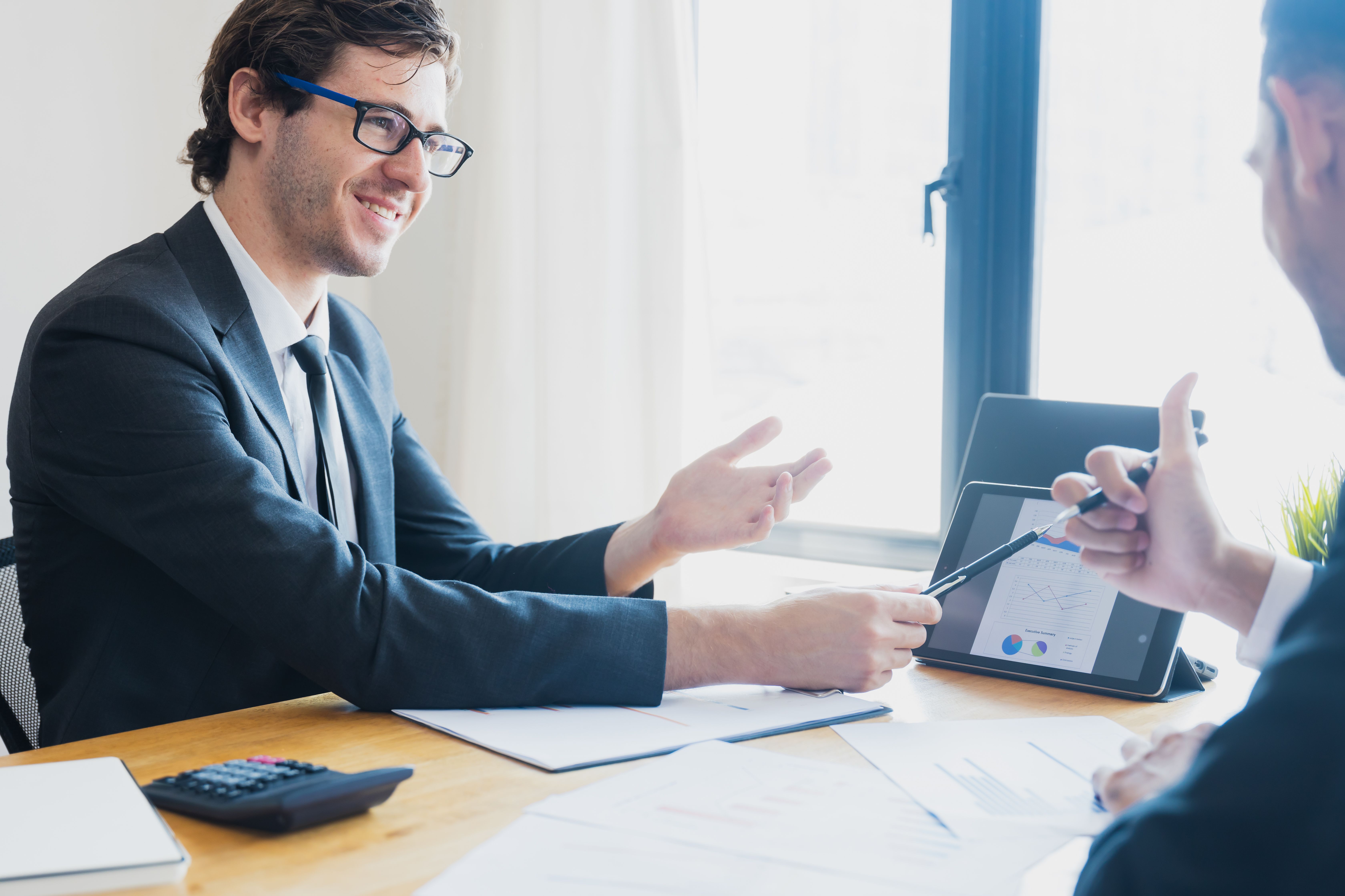 Two business professionals in suits discussing financial charts displayed on a tablet during a workplace meeting.